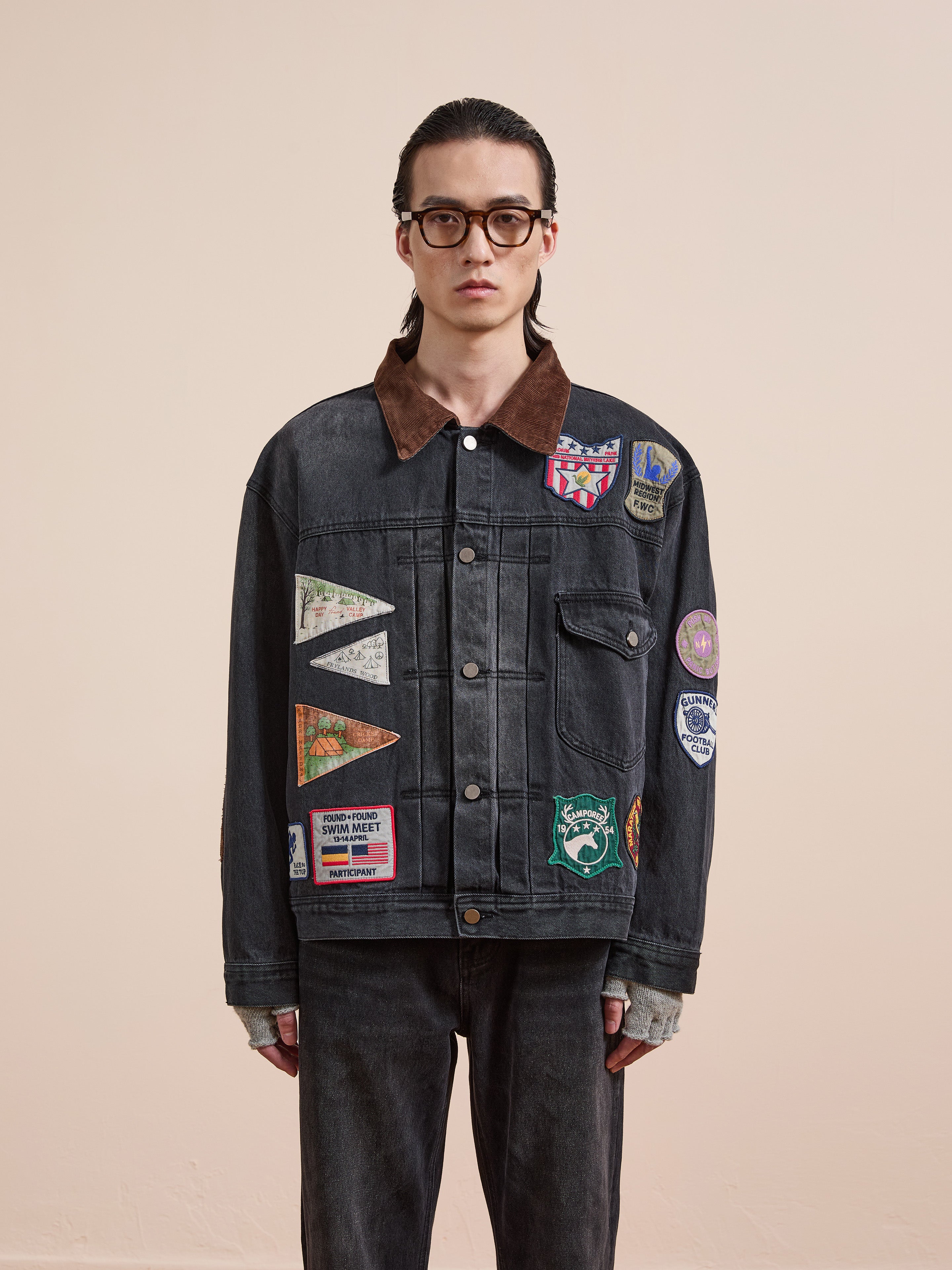 A person with long hair and glasses models the FOUND Denim Patchwork Jacket, featuring a corduroy collar and Americana patches, styled with dark pants against a plain light background.