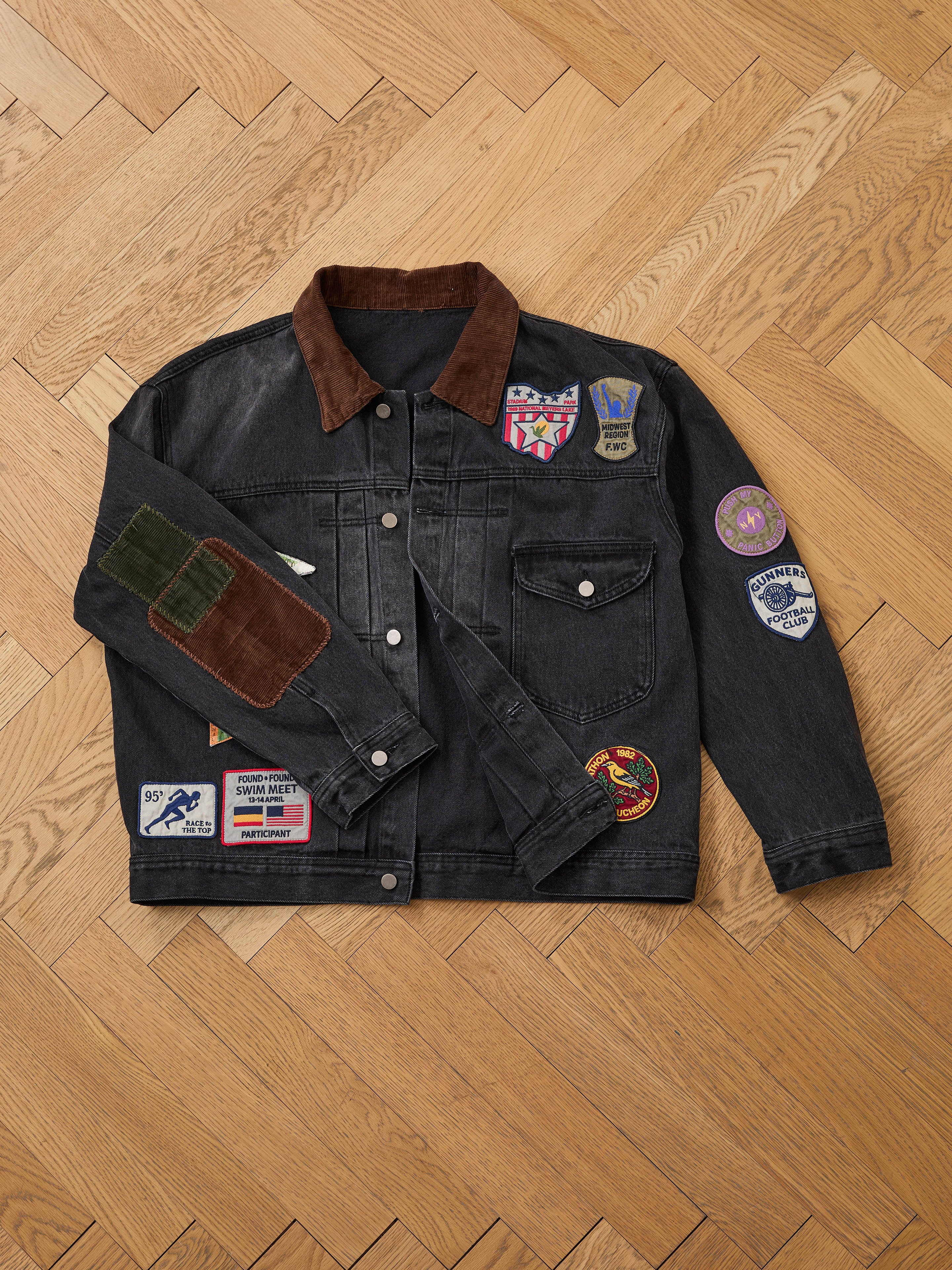 The FOUND Denim Patchwork Jacket, featuring vibrant Americana patches and a brown corduroy collar, is displayed flat on a wooden herringbone floor.