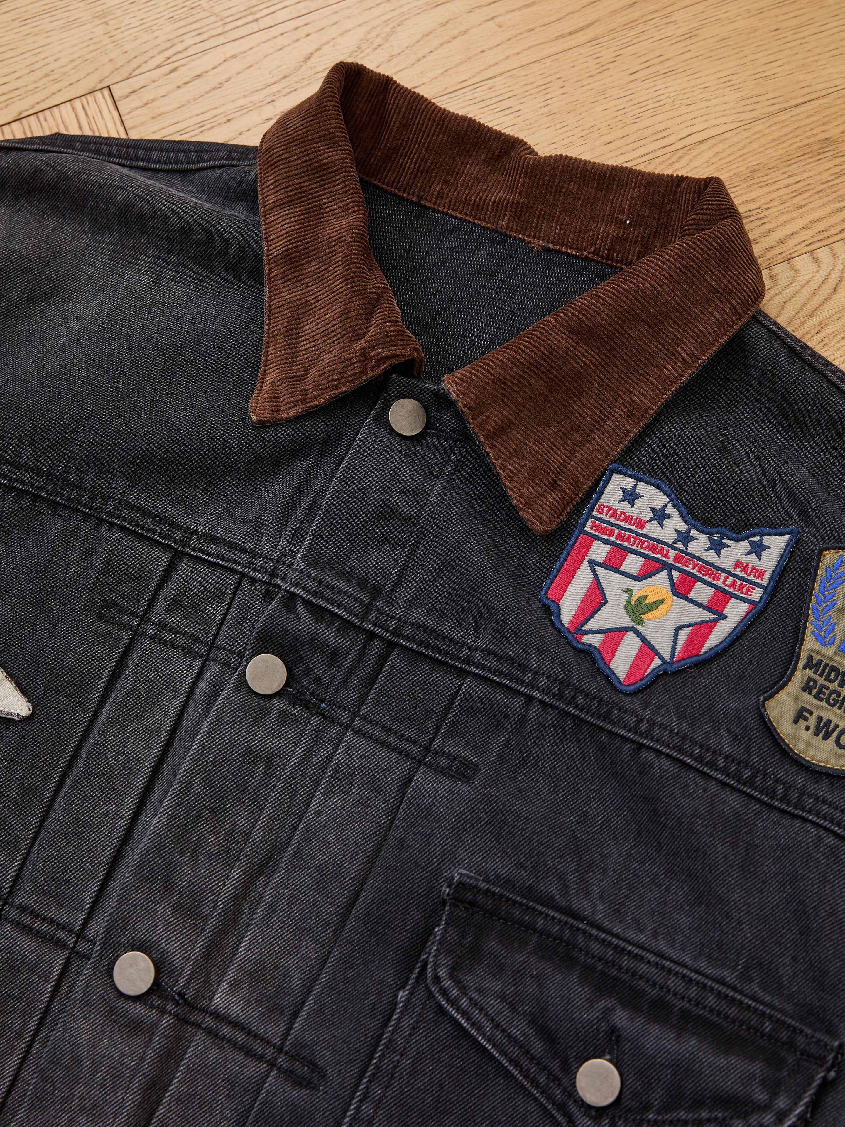 The FOUND Denim Patchwork Jacket features a brown corduroy collar, metal buttons, and Americana chest patches, shown laid flat on a wooden floor.