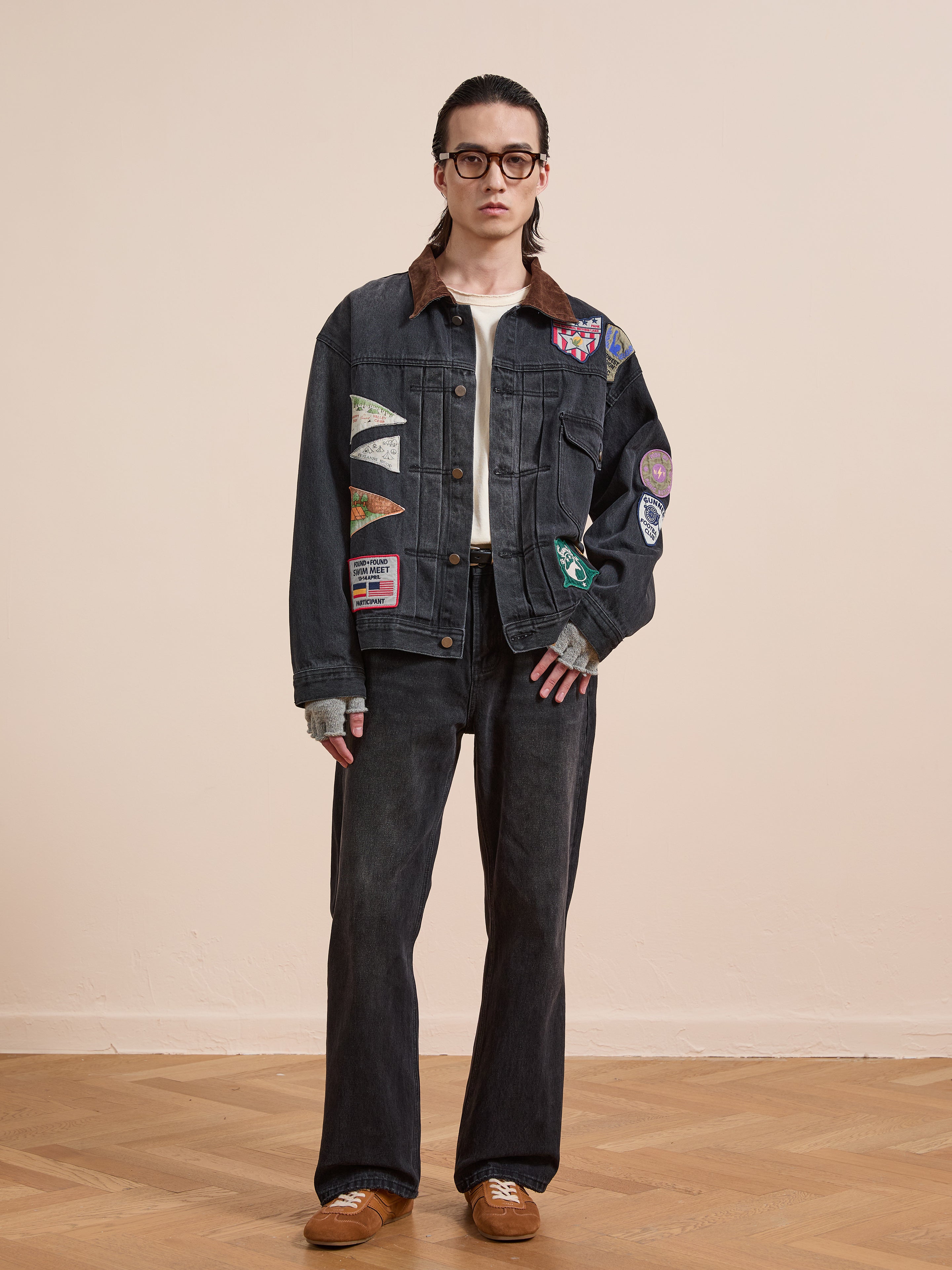 Person stands indoors against a light wall wearing the FOUND Denim Patchwork Jacket with Americana patches and a corduroy collar, paired with matching dark jeans, brown shoes, glasses, and slicked-back hair.