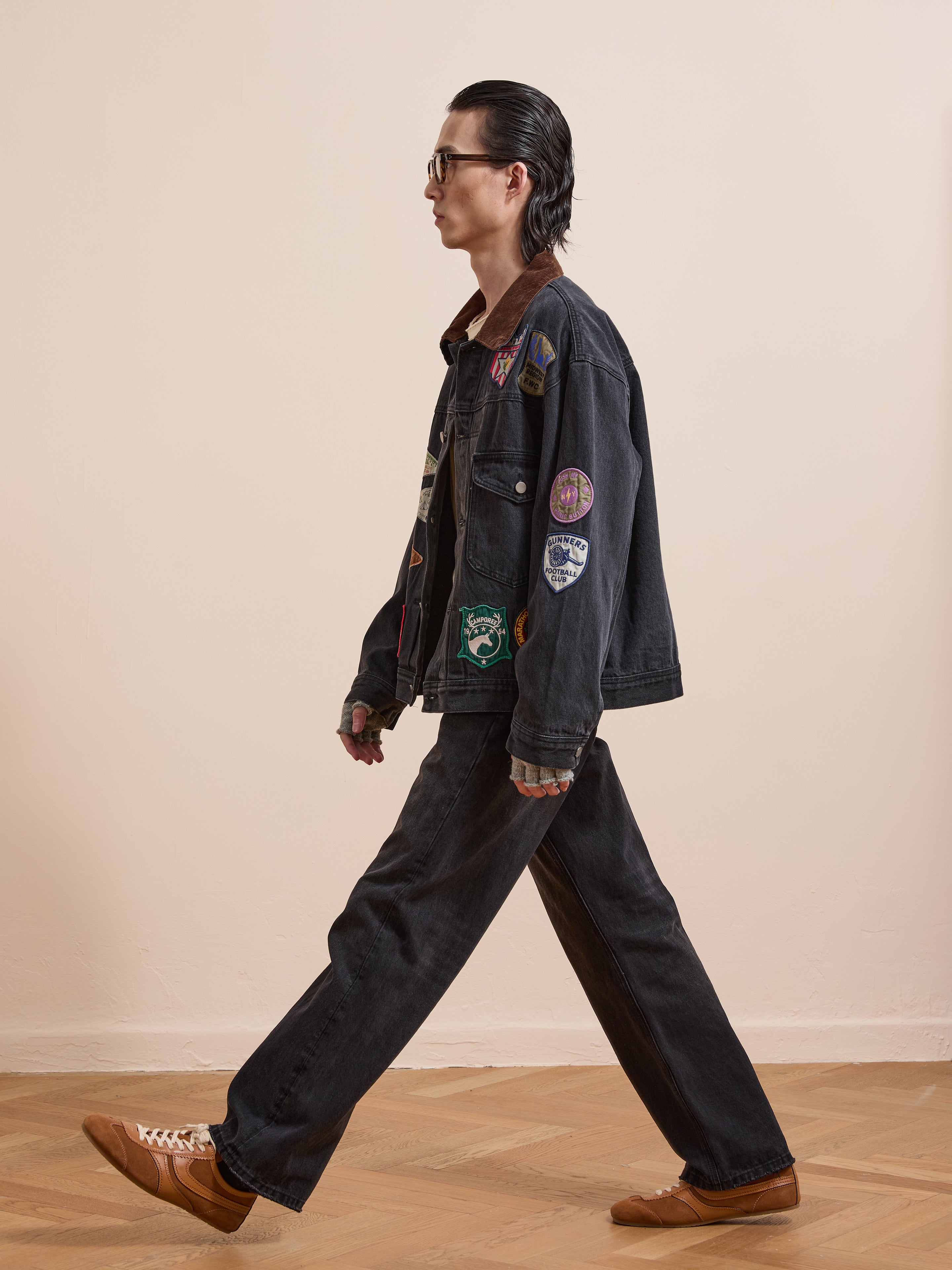 A person walks indoors in profile, wearing FOUND’s Denim Patchwork Jacket with Americana patches and a corduroy collar, matching wide-leg jeans, brown shoes, and glasses.