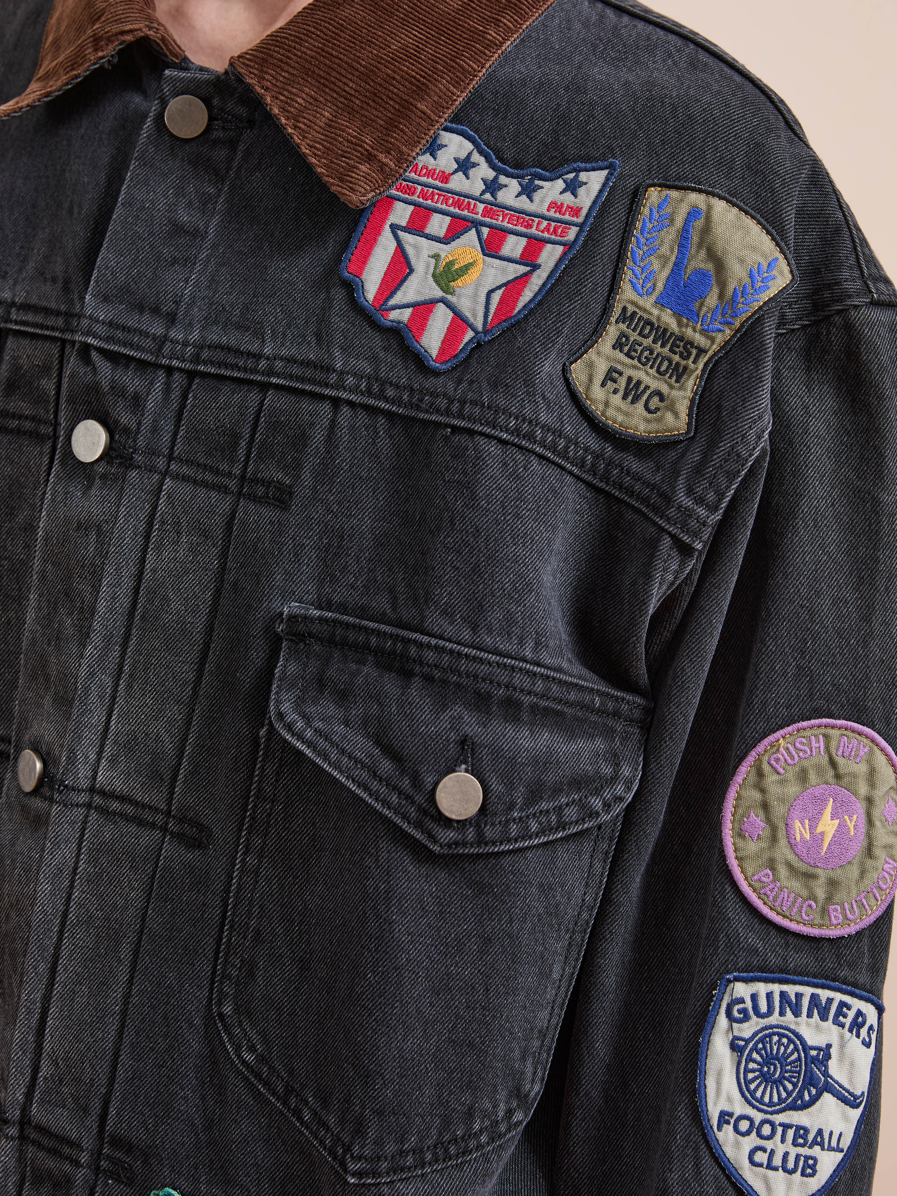 Close-up of the FOUND Denim Patchwork Jacket, featuring a corduroy collar and multiple colorful Americana patches—shield, eagle, football club, and circular motifs—on the chest and sleeve.