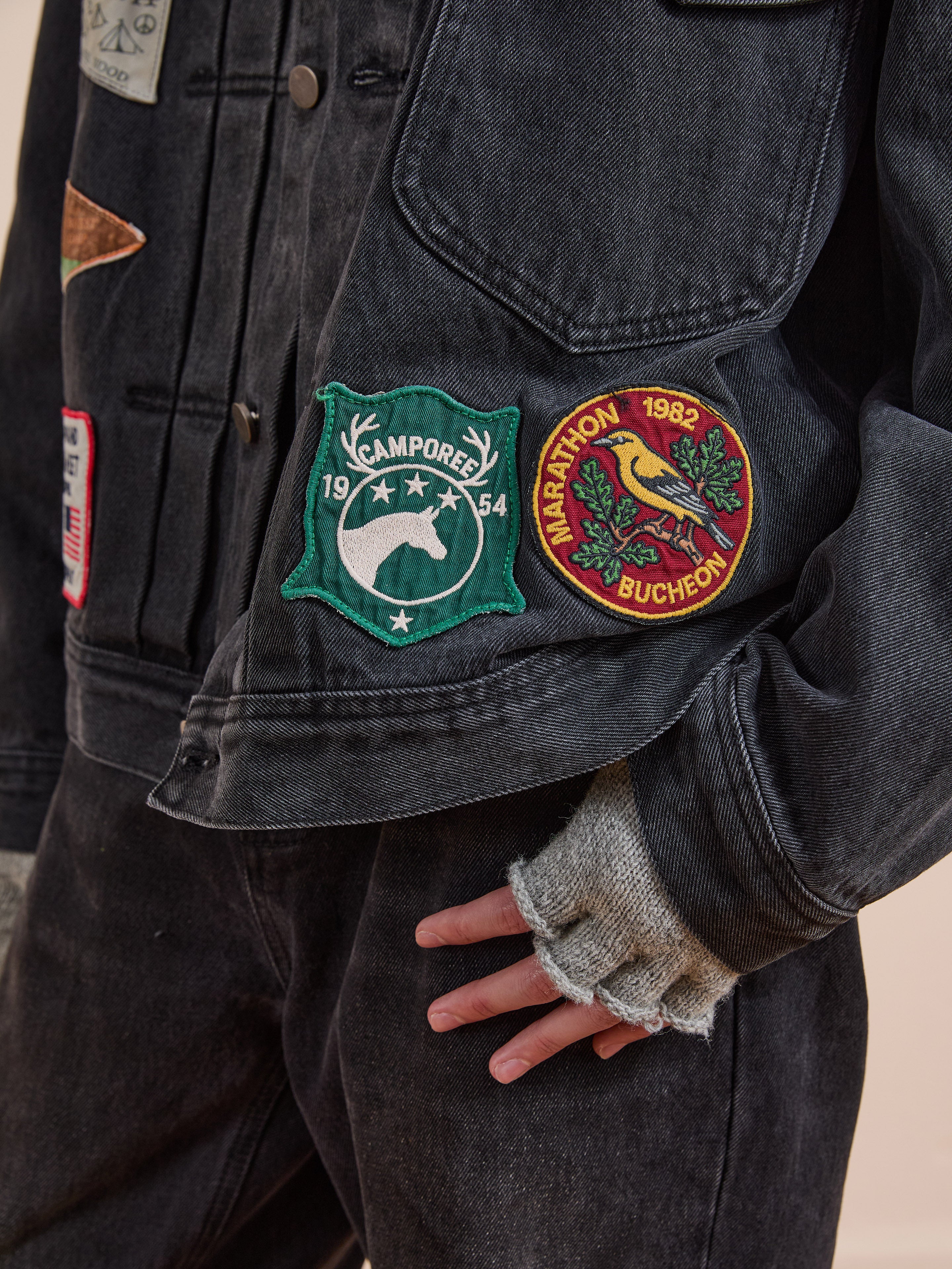 Someone wears FOUND’s Denim Patchwork Jacket—a black vintage denim piece with a corduroy collar, Americana patches—matching pants, and grey fingerless gloves.