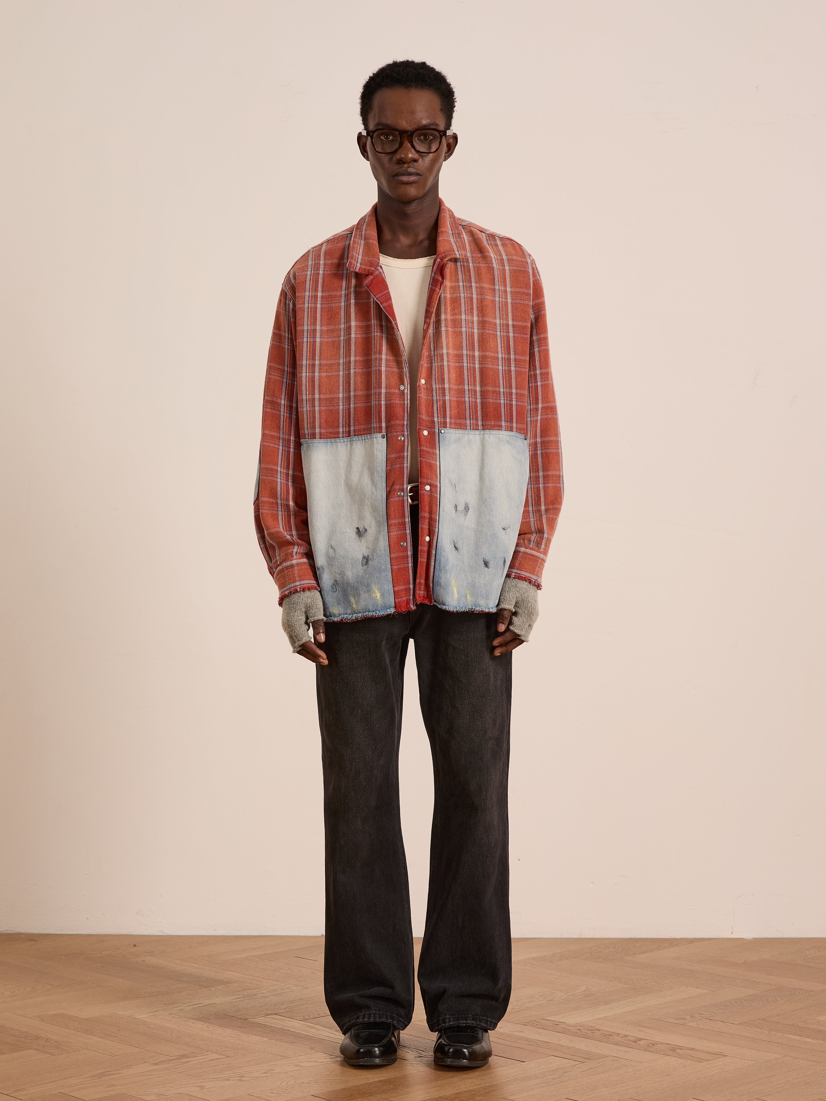 A person indoors wears the FOUND Plaid Denim Shirt with large carpenter pockets, glasses, a white undershirt, gray fingerless gloves, black jeans, and black shoes—showcasing a relaxed unisex workwear style.
