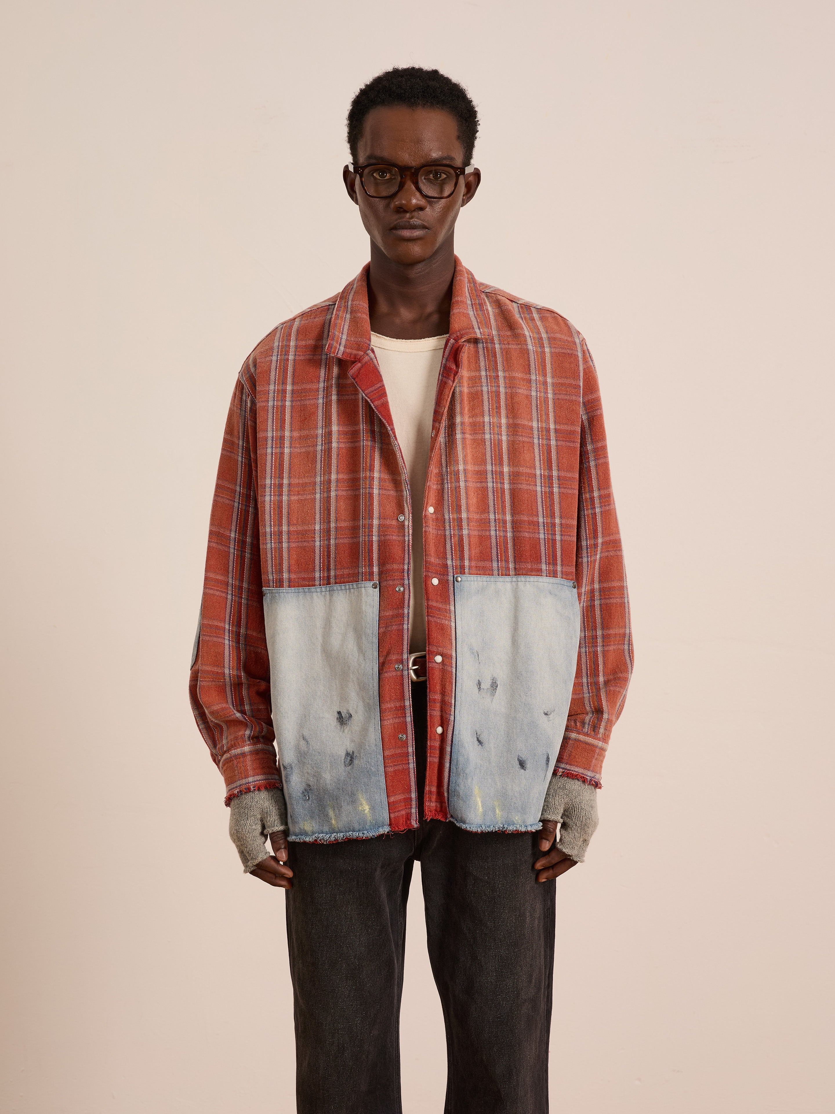 A man in glasses wears the FOUND Plaid Denim Shirt, featuring workwear-inspired red plaid and large denim carpenter pockets, paired with distressed fingerless gloves, a white undershirt, and dark pants against a plain background.
