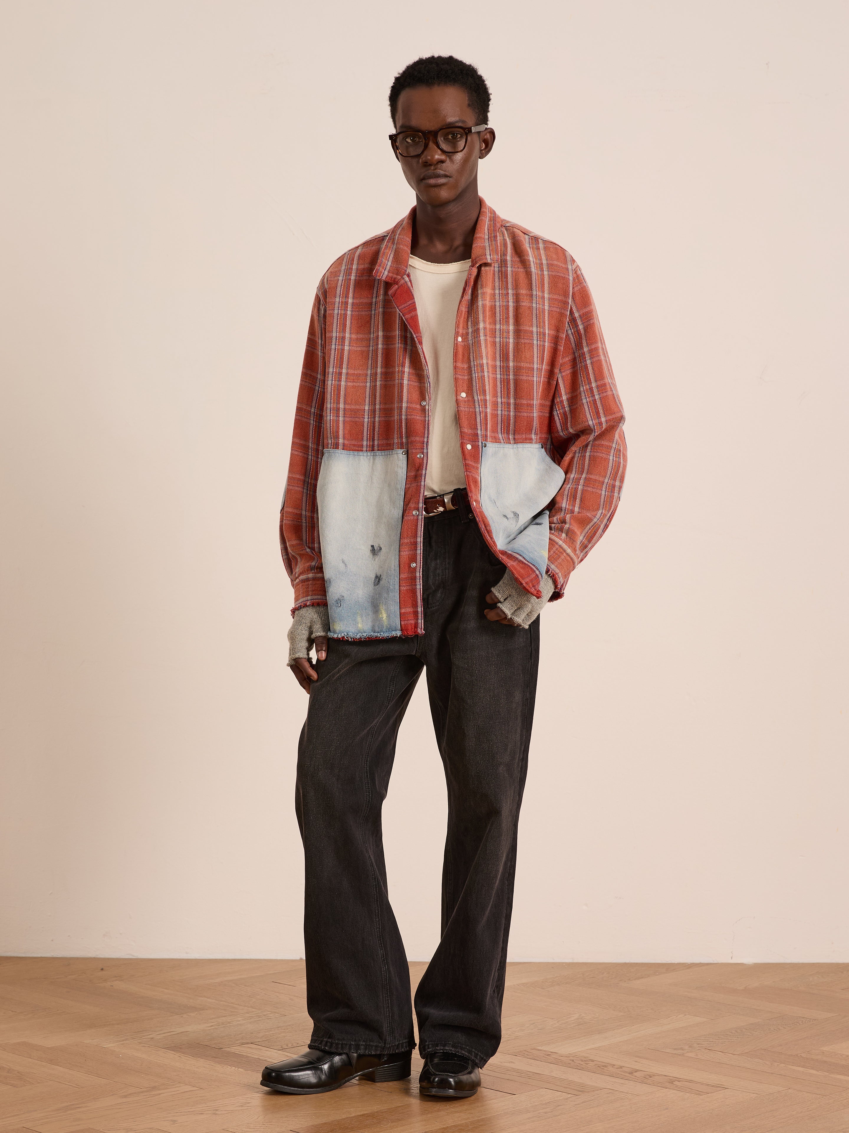 A person stands indoors against a plain background wearing the FOUND Plaid Denim Shirt—an orange unisex relaxed fit with carpenter pockets—paired with a white t-shirt, dark jeans, black shoes, and glasses.