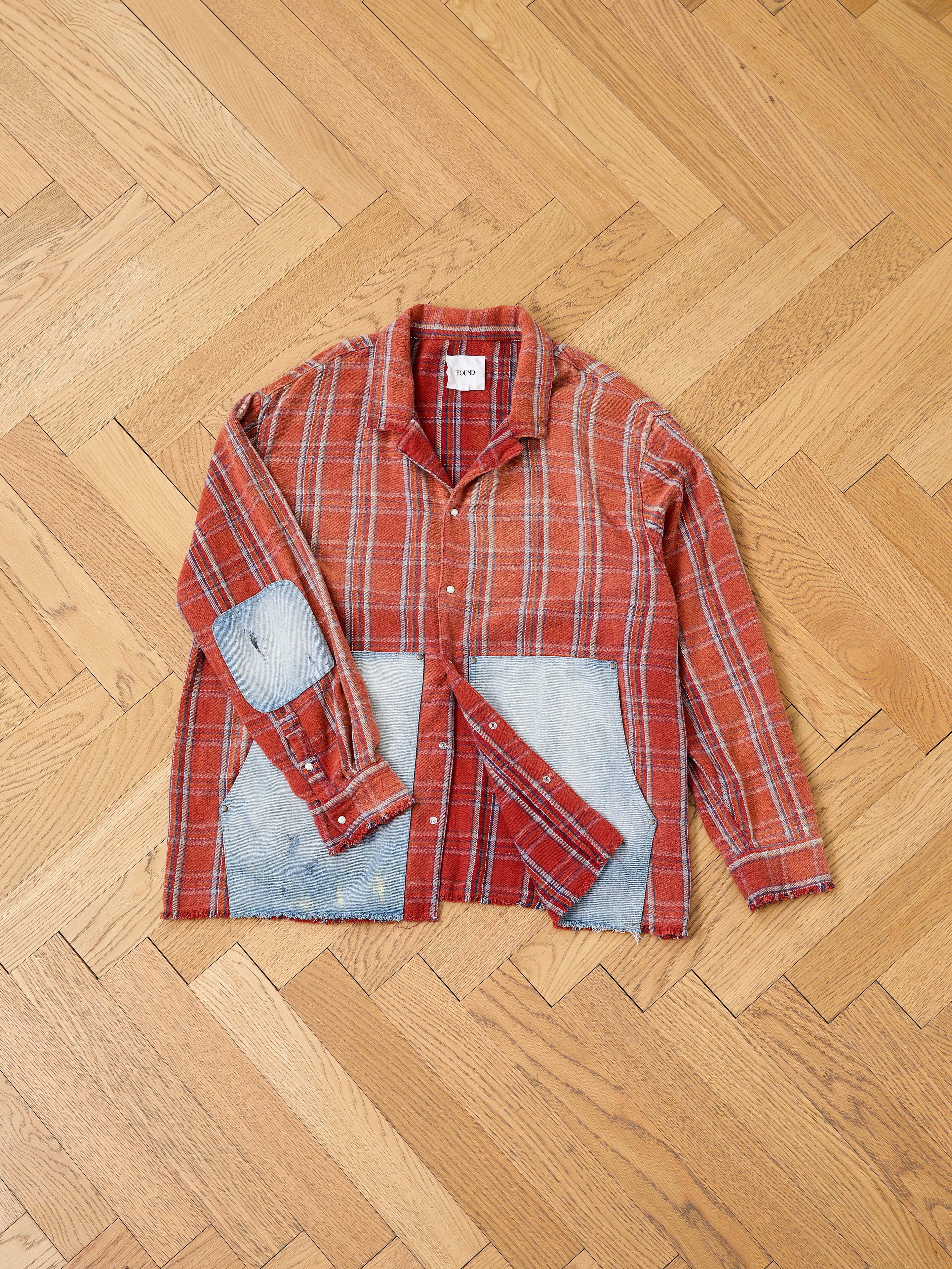 The FOUND Plaid Denim Shirt features red plaid fabric, denim carpenter pockets, and patchwork on the elbows and lower front, shown laid flat on a wooden herringbone floorâunisex workwear style.