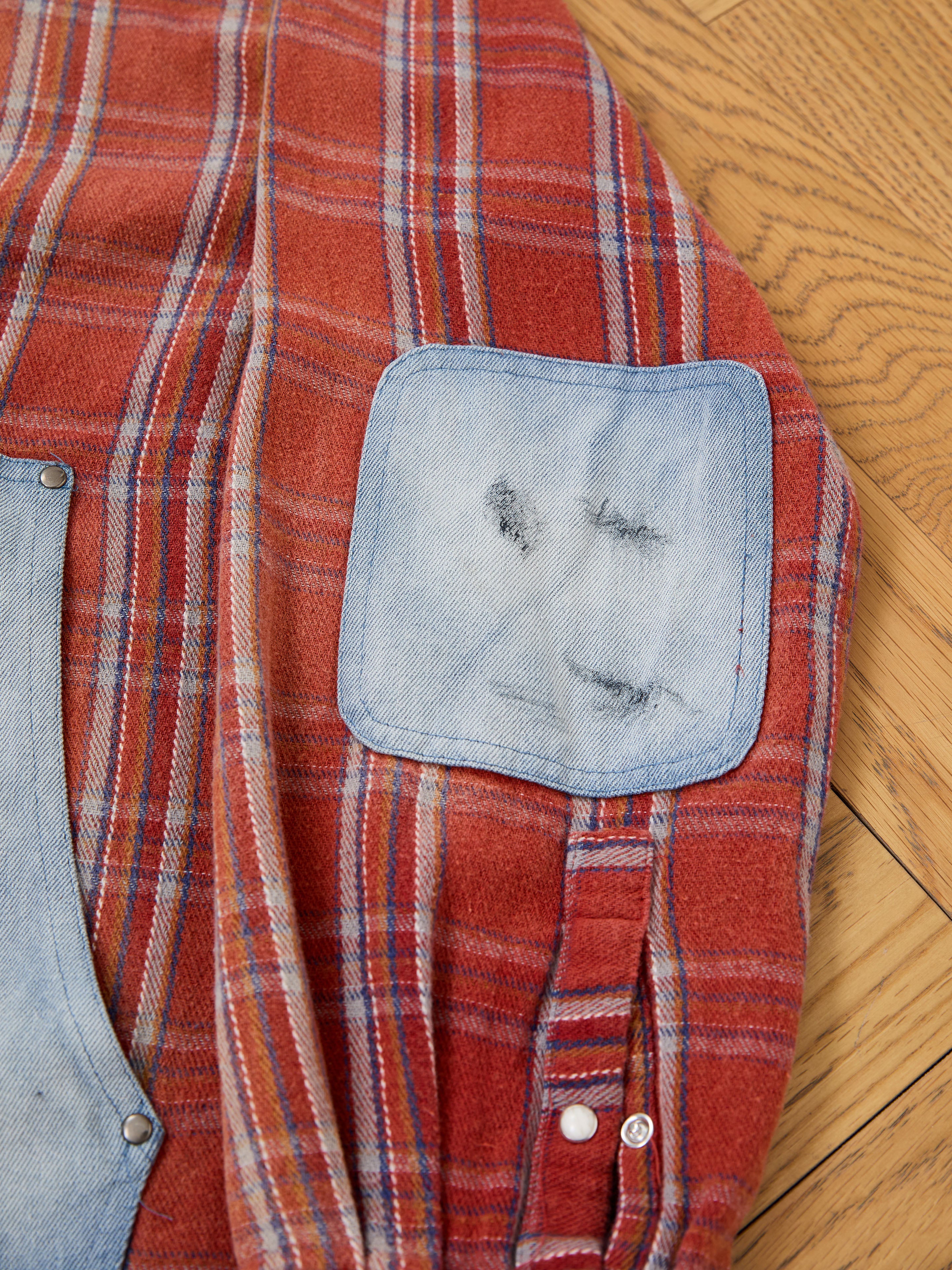 The FOUND Plaid Denim Shirt features a red plaid design with a light blue denim elbow patch showing wear and black marks, capturing classic unisex workwear style. Photographed on a wooden floor.