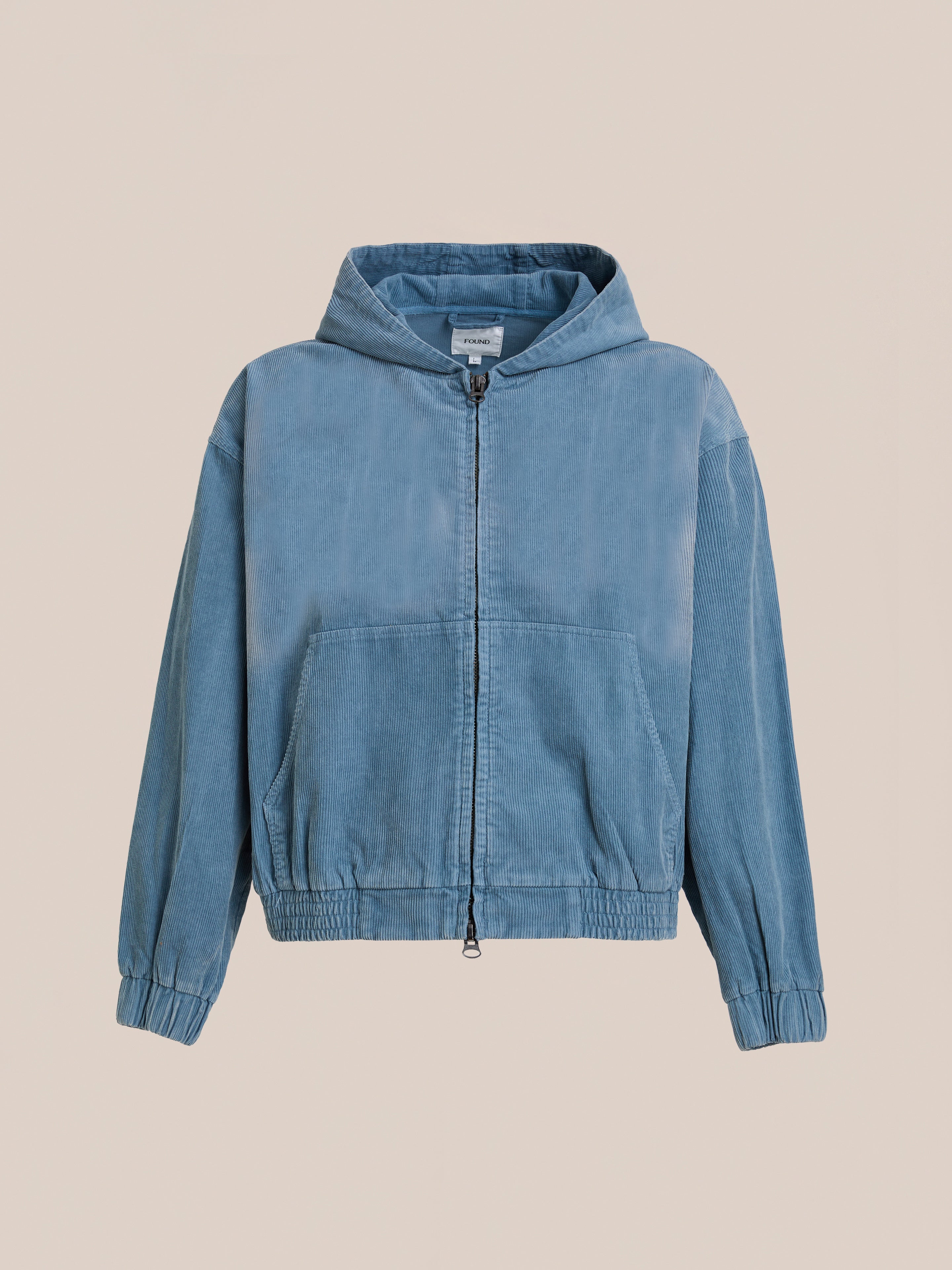 The FOUND Corduroy Zip Up Hoodie in light blue features front pockets, elastic cuffs, and waistband, shown against a plain beige background.