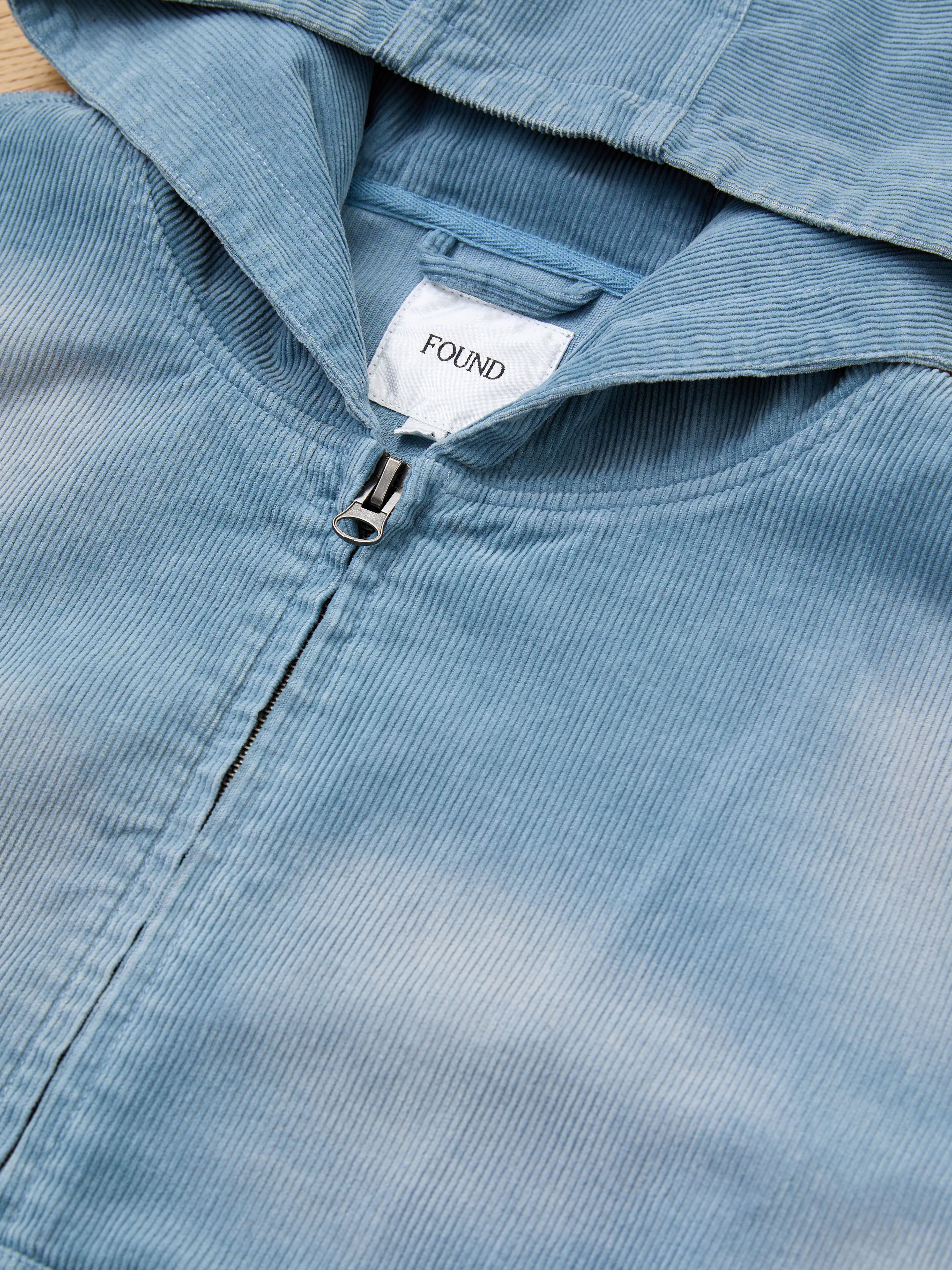 Close-up of the Corduroy Zip Up Hoodie by FOUND, shown in faded blue with a zipper and hood, and a white "FOUND" label inside the collar.