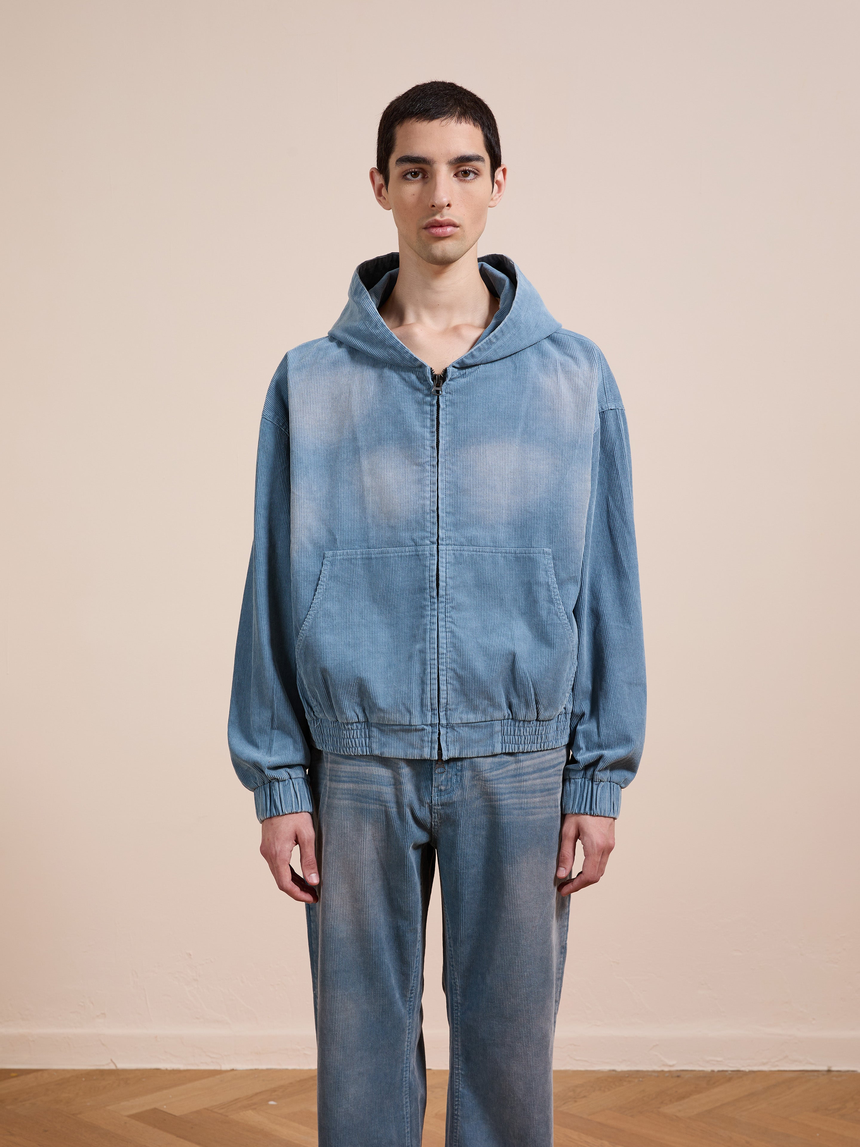 A person stands indoors facing forward against a plain beige background, wearing the FOUND Corduroy Zip Up Hoodie in faded blue with matching denim pants.