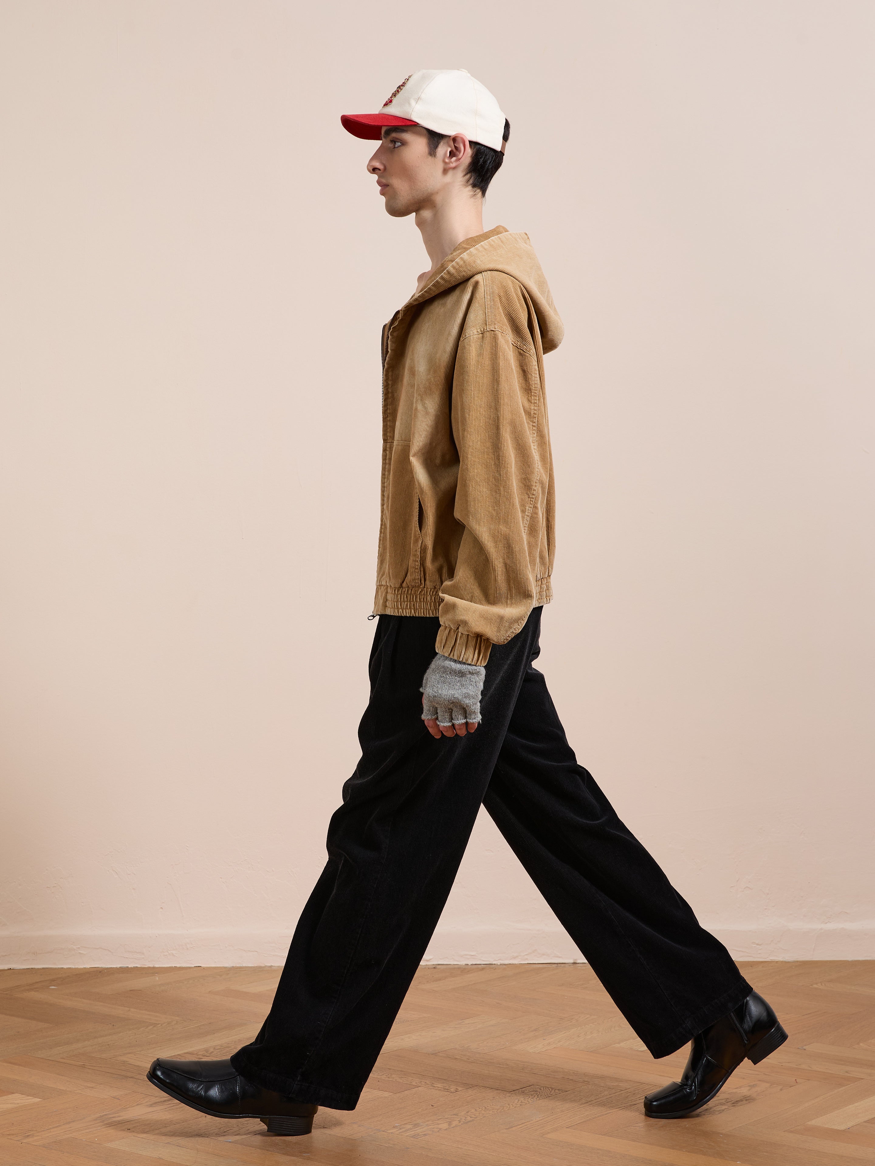 A person in FOUND’s Corduroy Zip Up Hoodie, black pants, black shoes, gray gloves, and a white-and-red cap walks on a wooden floor against a plain beige wall.