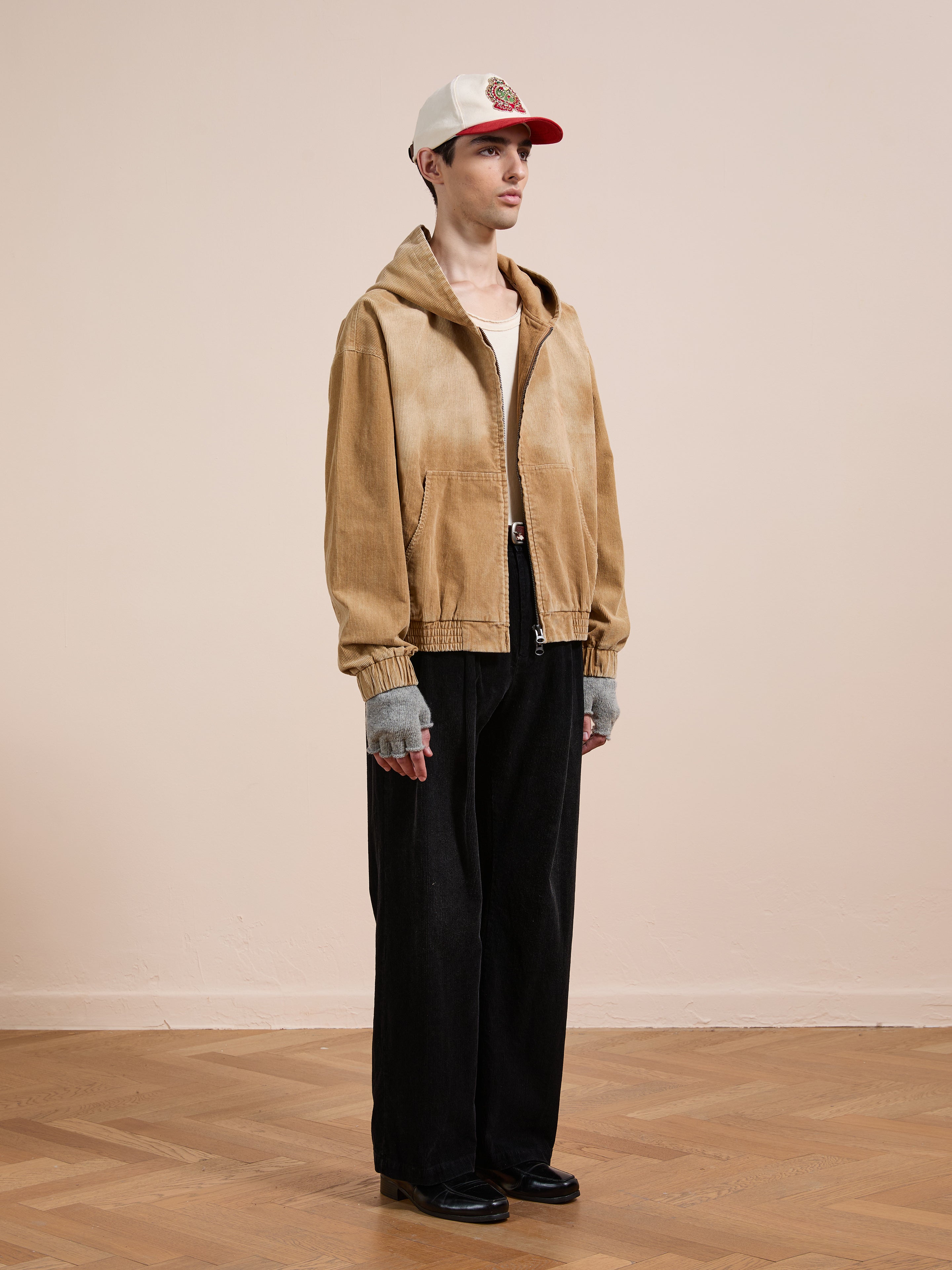 Indoors, a person wears the FOUND Corduroy Zip Up Hoodie in tan with black wide-leg pants, black shoes, gray fingerless gloves, and a white and red baseball cap for a vintage-inspired look.