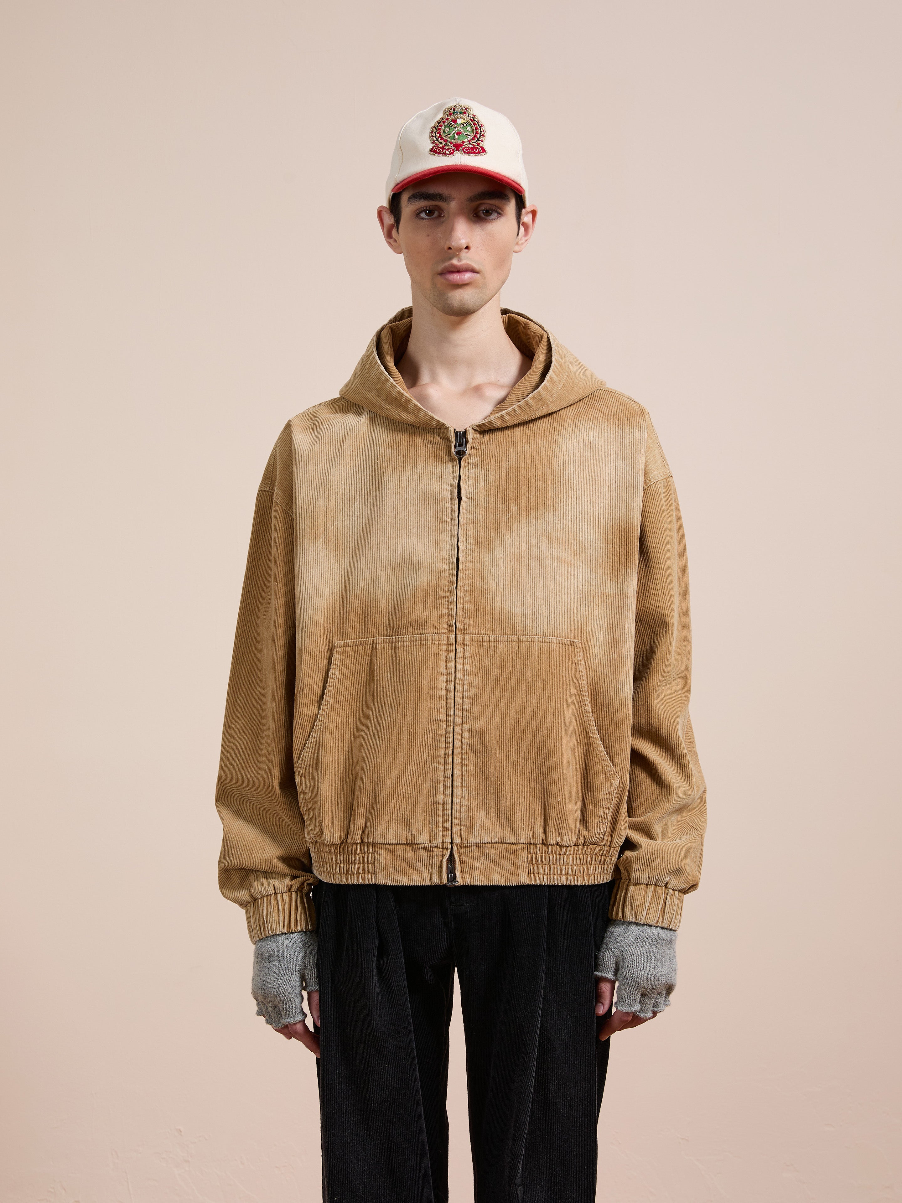 A person in the FOUND Corduroy Zip Up Hoodie (tan, unisex relaxed fit), black pants, gray fingerless gloves, and a white cap with a red brim stands before a plain beige background.