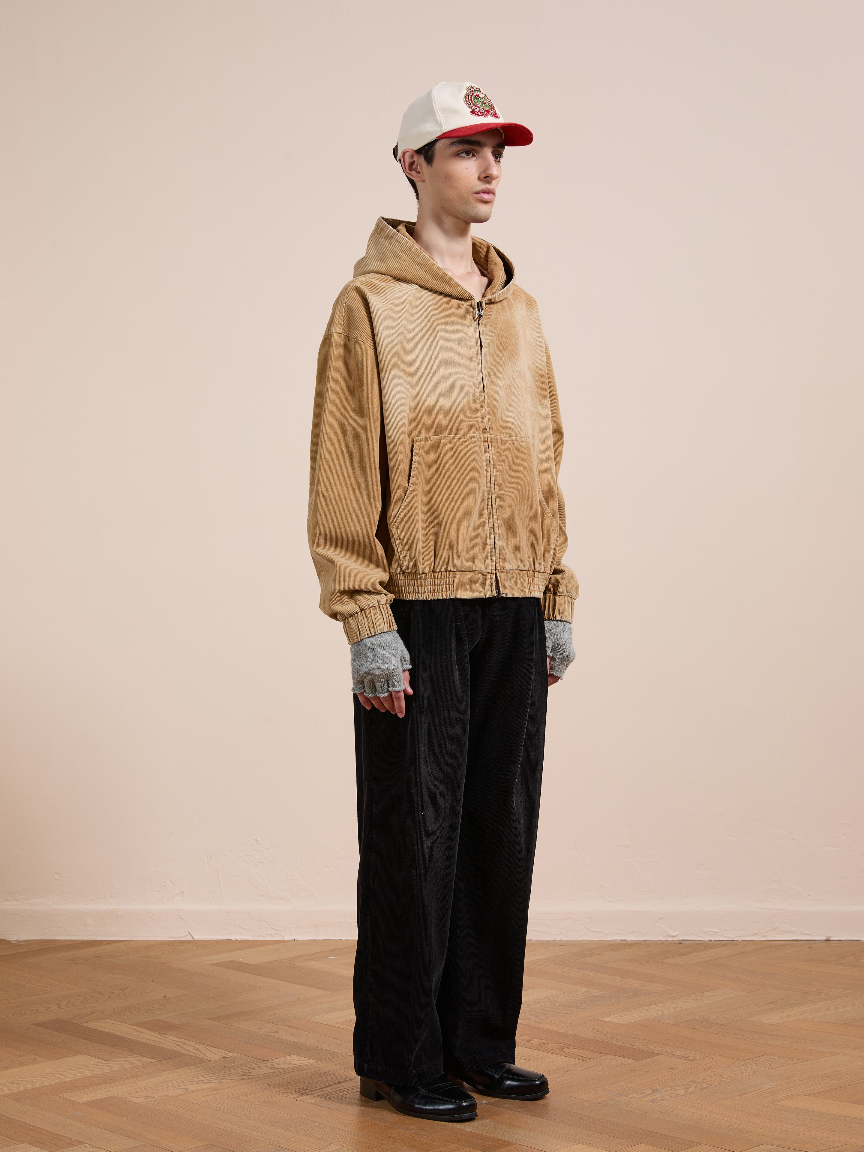 A person indoors wears the FOUND Corduroy Zip Up Hoodie in a vintage beige, unisex relaxed fit, styled with black pants, gray fingerless gloves, black shoes, and a white and red baseball cap against a plain background.