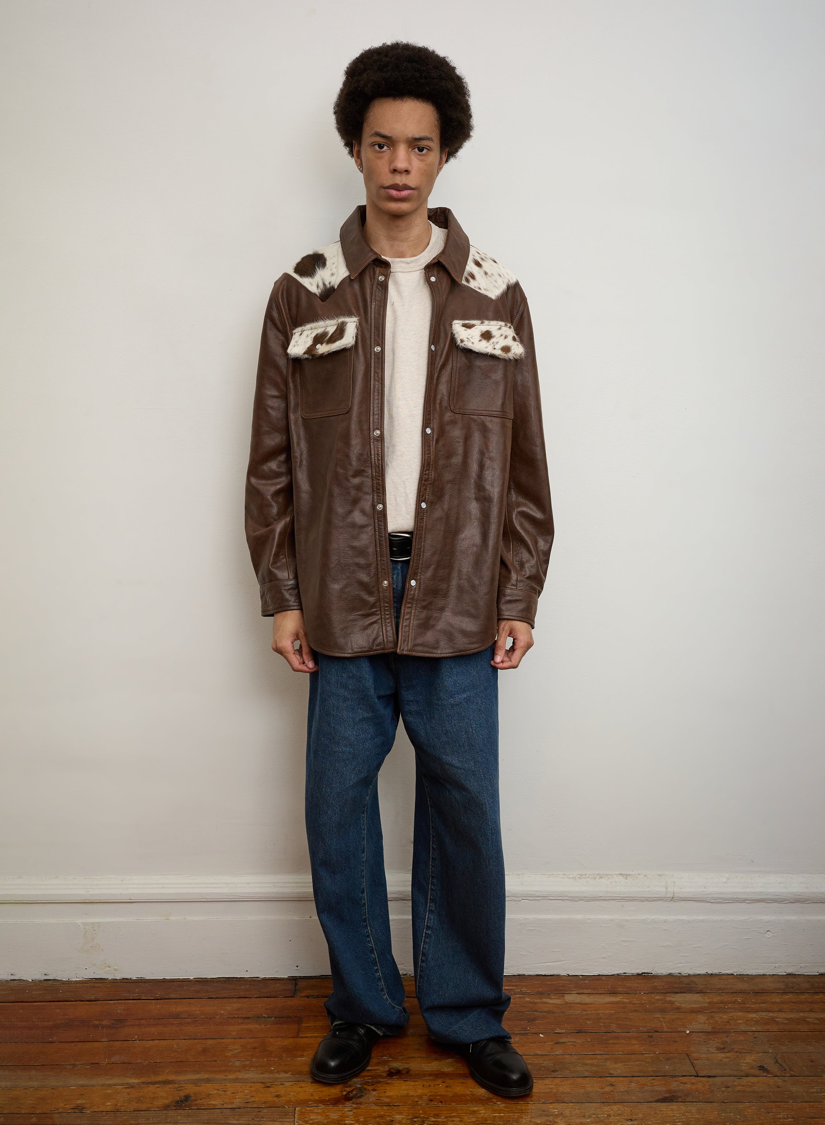 A person stands indoors against a light wall and wooden floor, wearing the FOUND Distressed Leather Cowhide Contrast Western Shirt with white fur patches, a beige shirt, blue jeans, and black shoes.