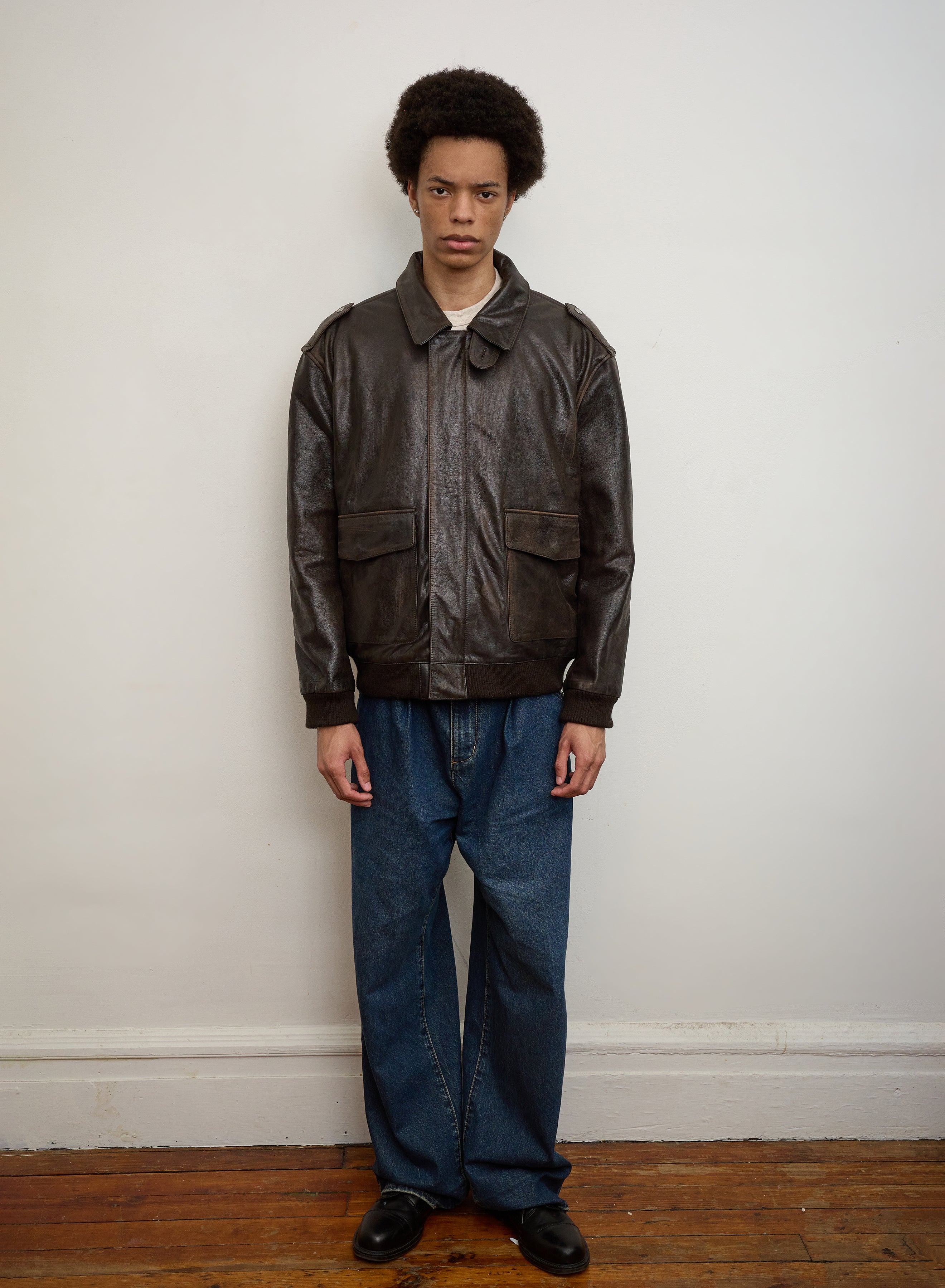 A person with short curly hair stands against a white wall, wearing the FOUND Distressed Leather Flight Jacket, blue jeans, and black shoes on a wooden floor.