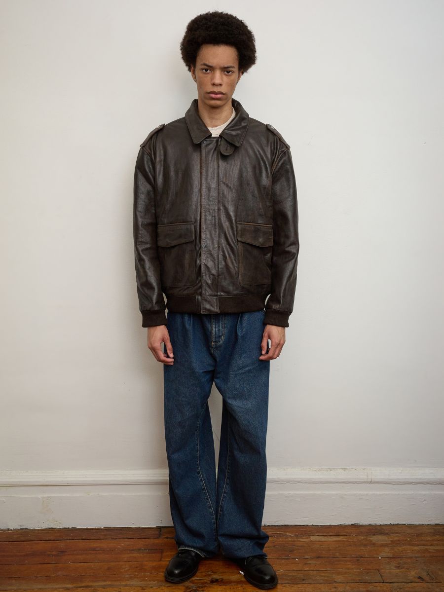 A person stands indoors against a plain white wall, wearing the FOUND Distressed Leather Flight Jacket, loose blue jeans, and black shoes, with a neutral expression.
