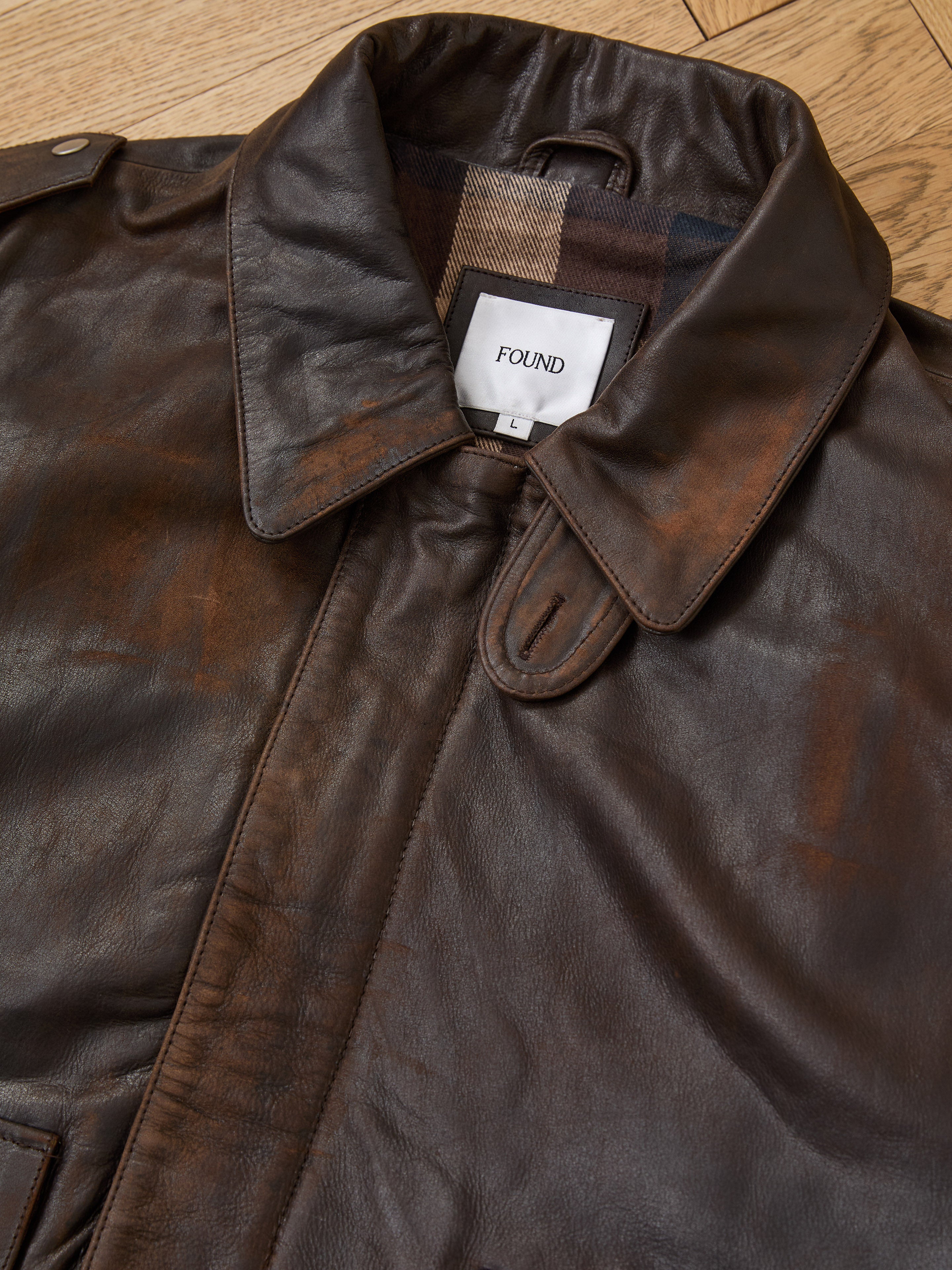 A close-up of the FOUND Distressed Leather Flight Jacket, a unisex brown genuine leather piece with collar and epaulettes, is shown laid flat on a wooden surface. The inside label reads "FOUND.