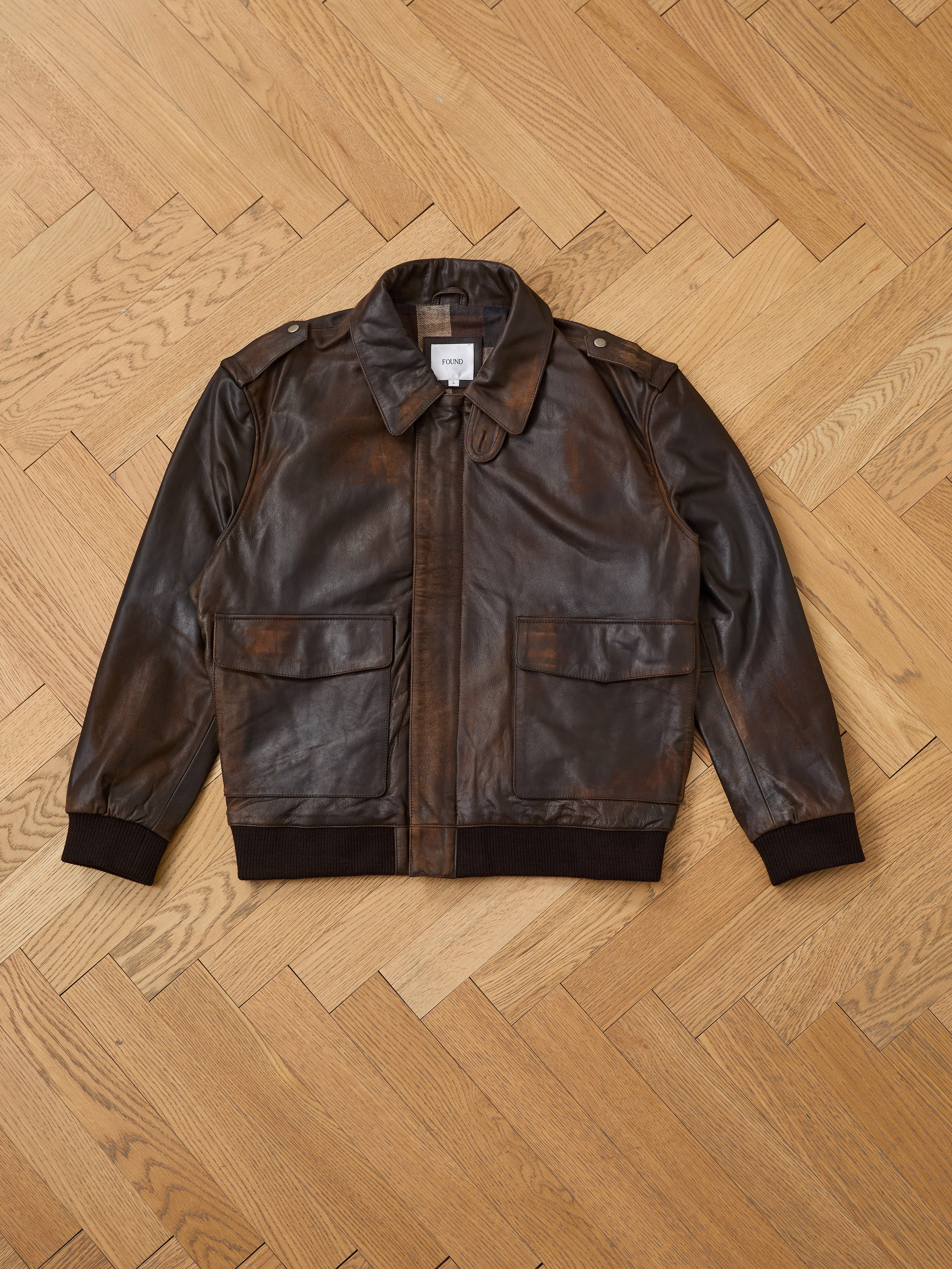 The FOUND Distressed Leather Flight Jacket, a stylish unisex piece with front flap pockets, black cuffs, and collar, is shown flat on a wooden herringbone floor—perfect for any wardrobe.
