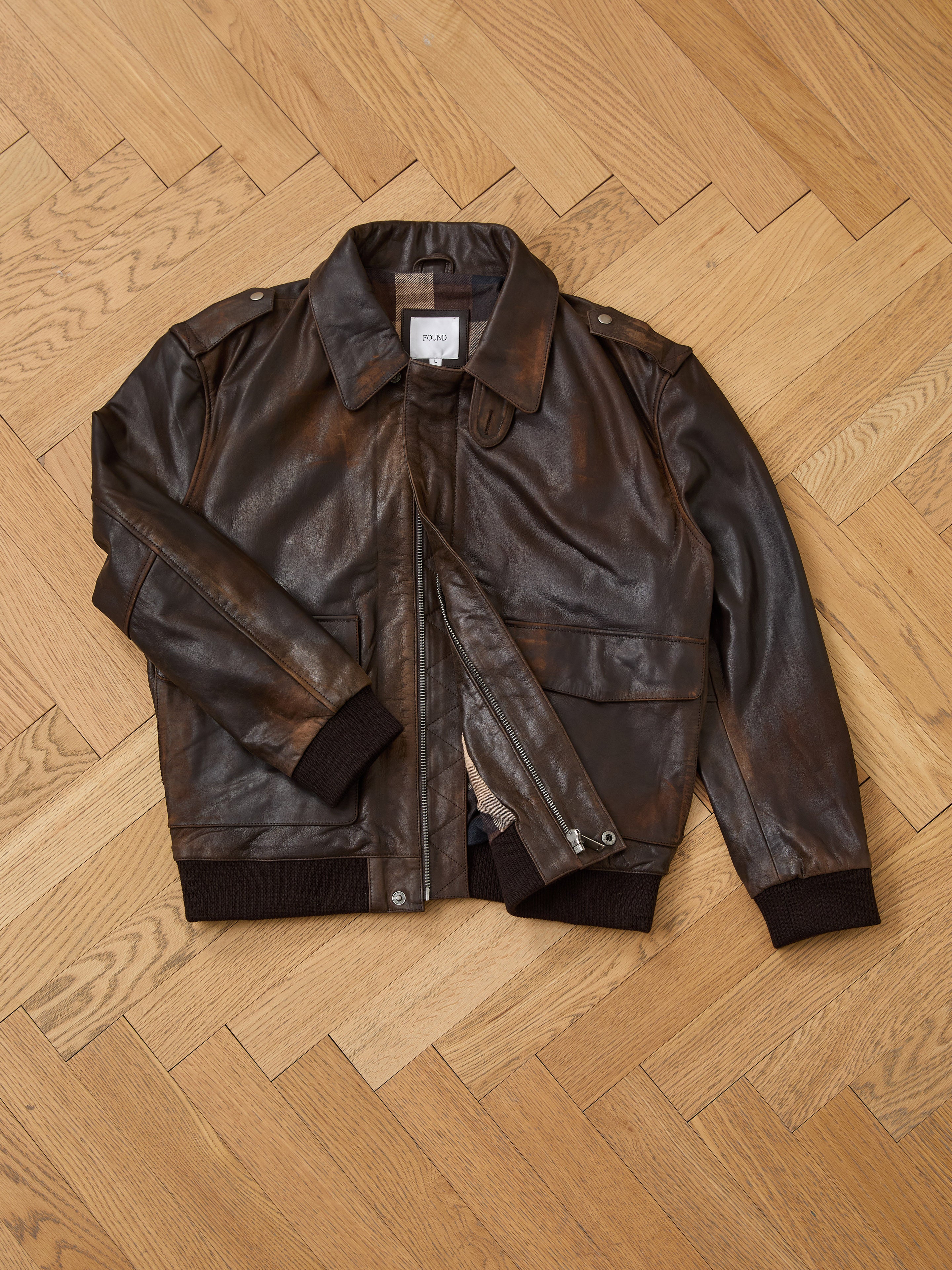 The FOUND Distressed Leather Flight Jacket, a classic unisex style with collar and front zipper, is displayed flat on a light herringbone wood floor.