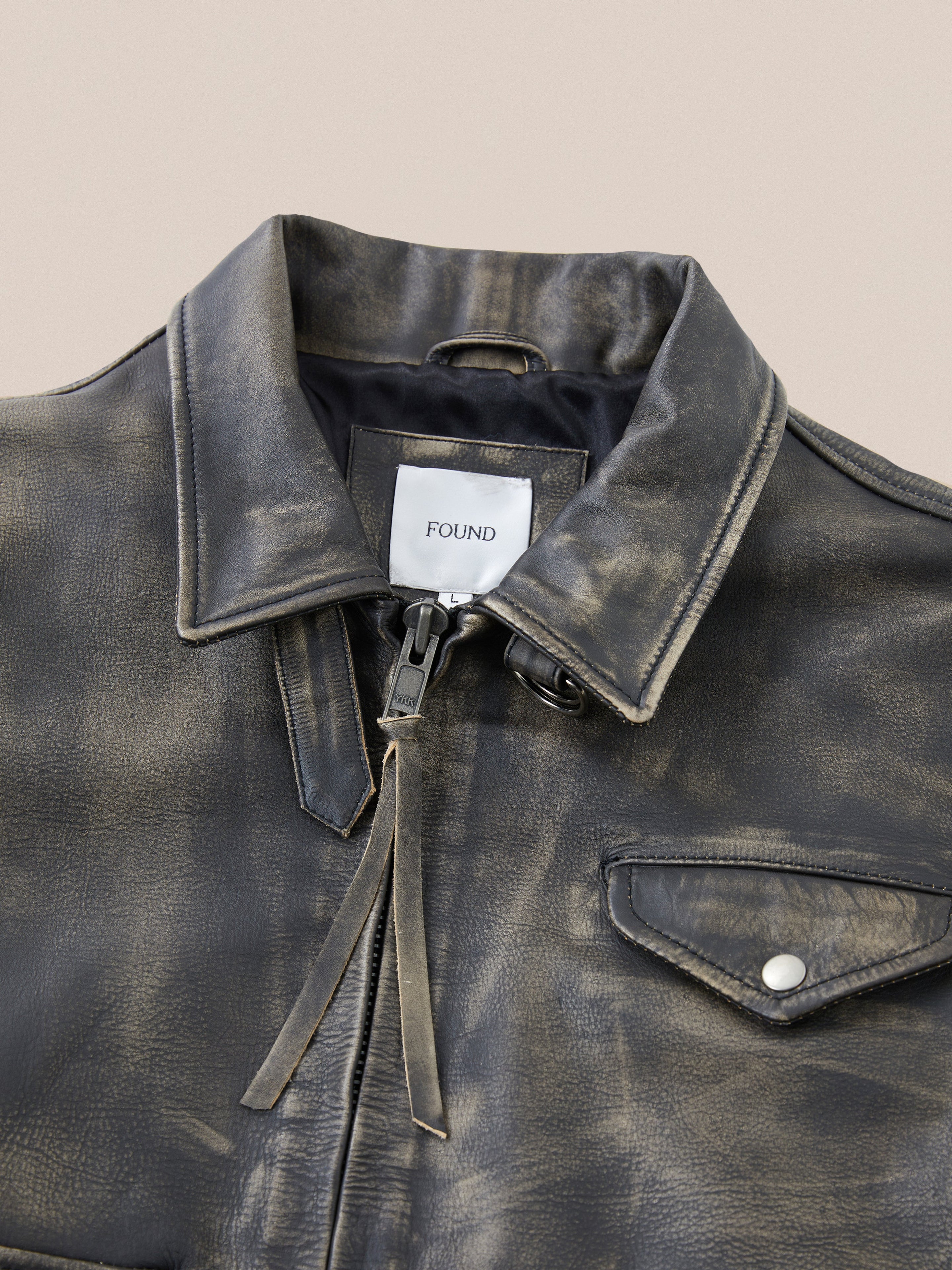 A distressed black cowhide leather jacket from Found, featuring a silver zipper, pointed collar, and buttoned chest pocket. Its antique feel and "Found" label enhance its unique charm.
