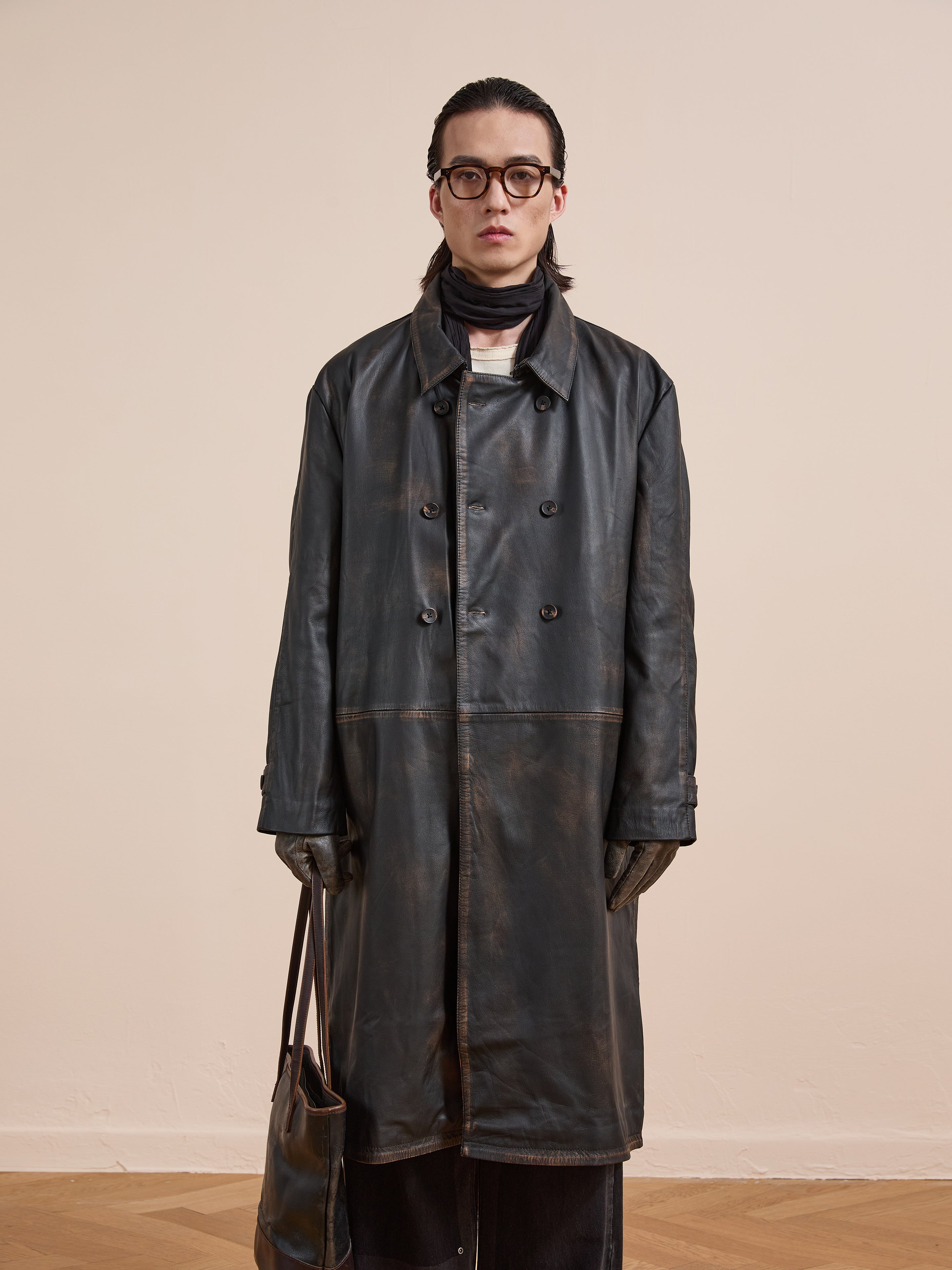 A person wears the FOUND Distressed Leather Trench Coat with glasses, gloves, scarf, and a matching leather bag, standing against a light beige wall and wooden floor.