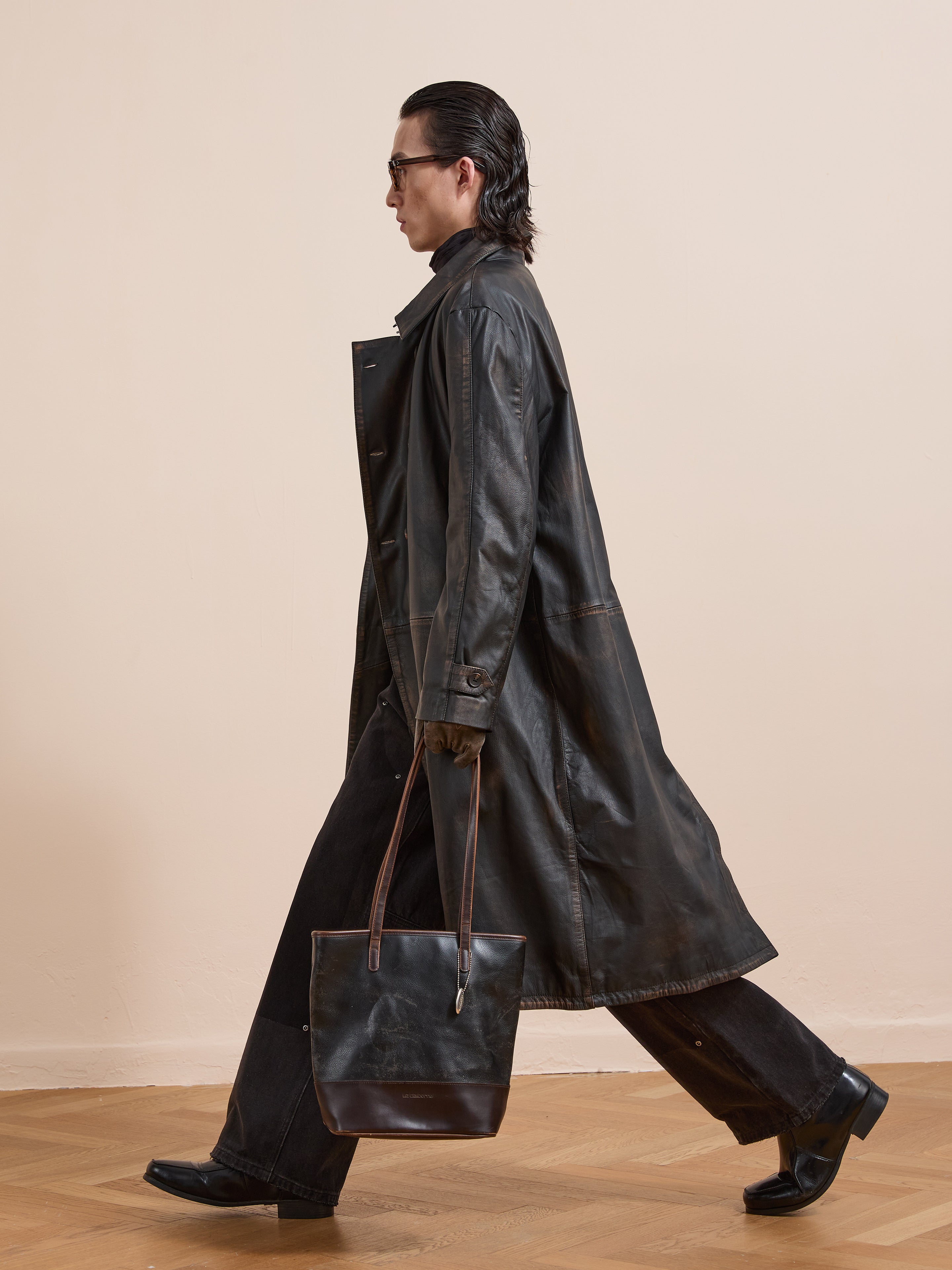 A person in FOUND's Distressed Leather Trench Coat, paired with black pants, boots, and a matching black leather tote, walks indoors.