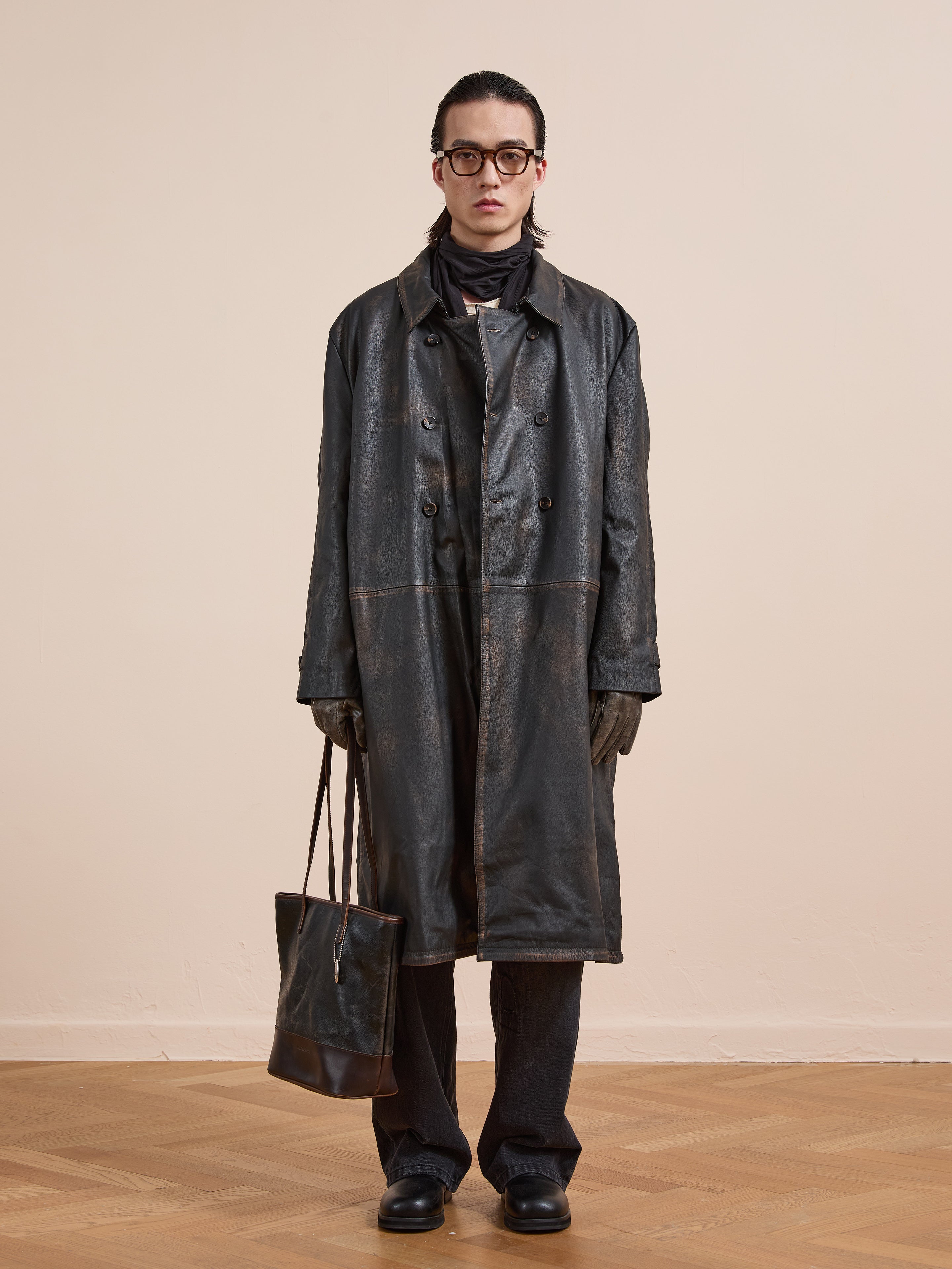 Indoors, a person wears the FOUND Distressed Leather Trench Coat with dark pants, black shoes, gloves, glasses, and holds a brown leather bag.