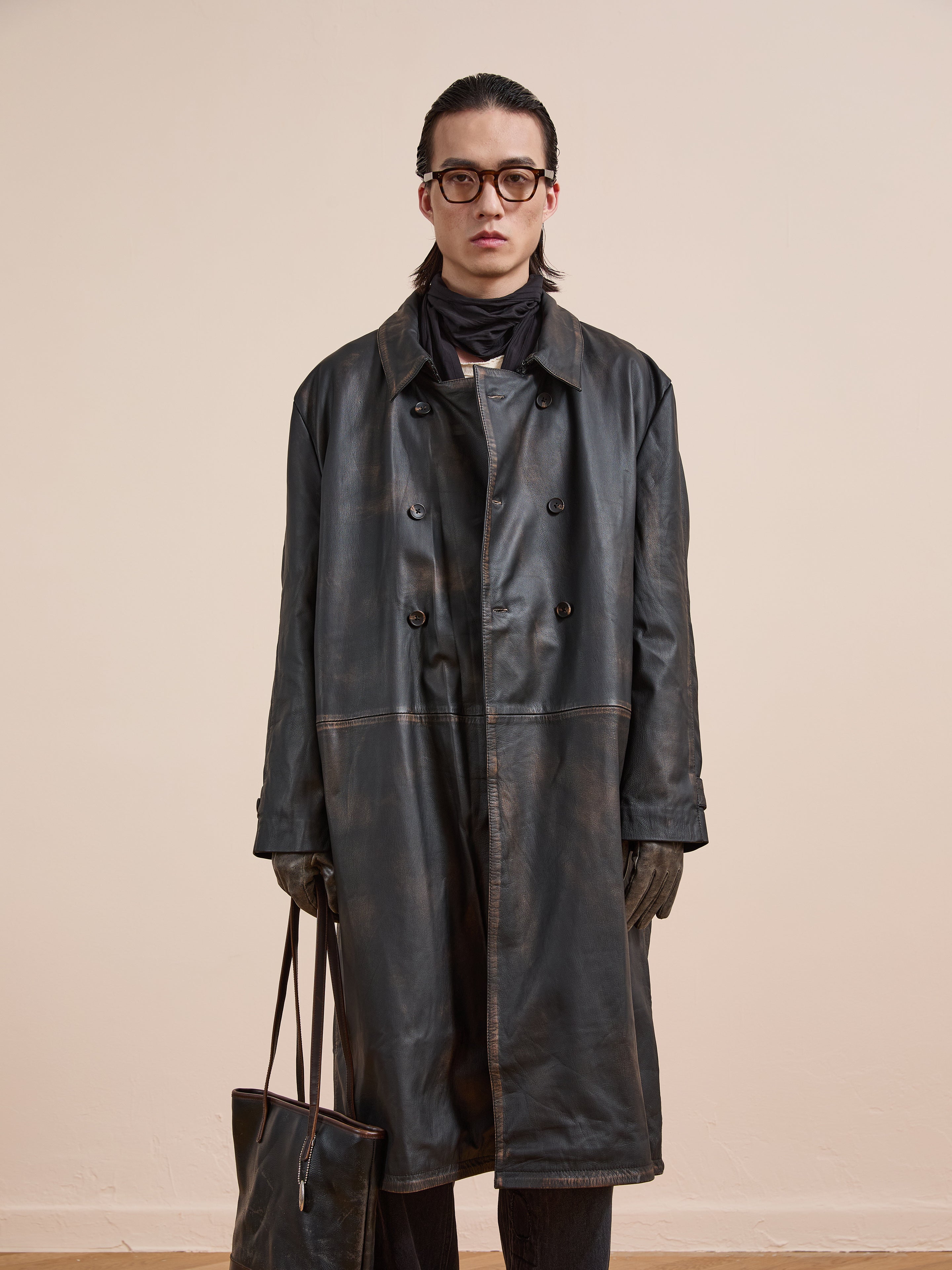A person in a FOUND Distressed Leather Trench Coat, black gloves, round glasses, and scarf holds a matching leather bag while standing against a plain beige background.