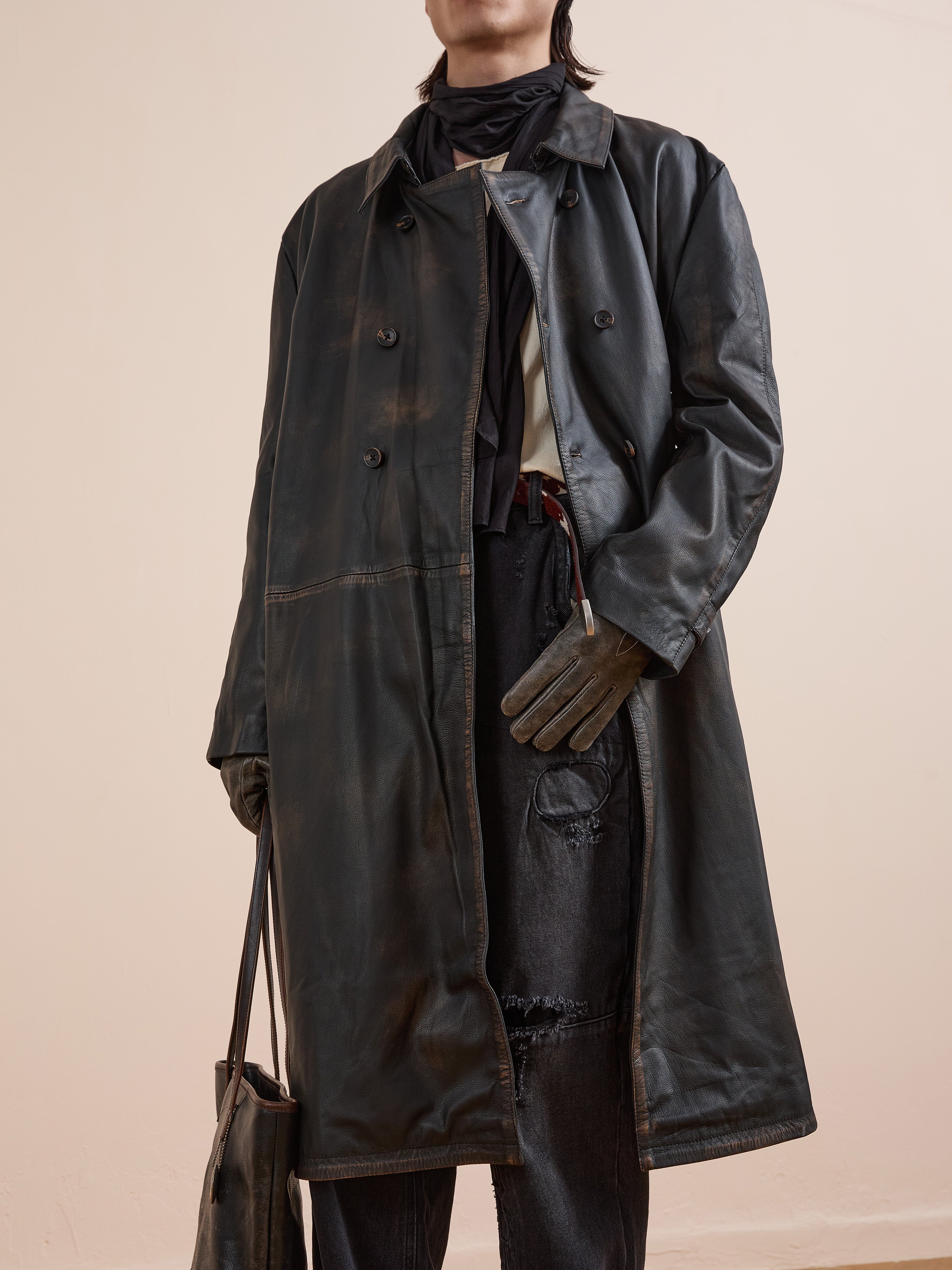 A person in the FOUND Distressed Leather Trench Coat, dark gloves, a black scarf, and ripped black jeans holds a matching leather bag. Their face is not visible.