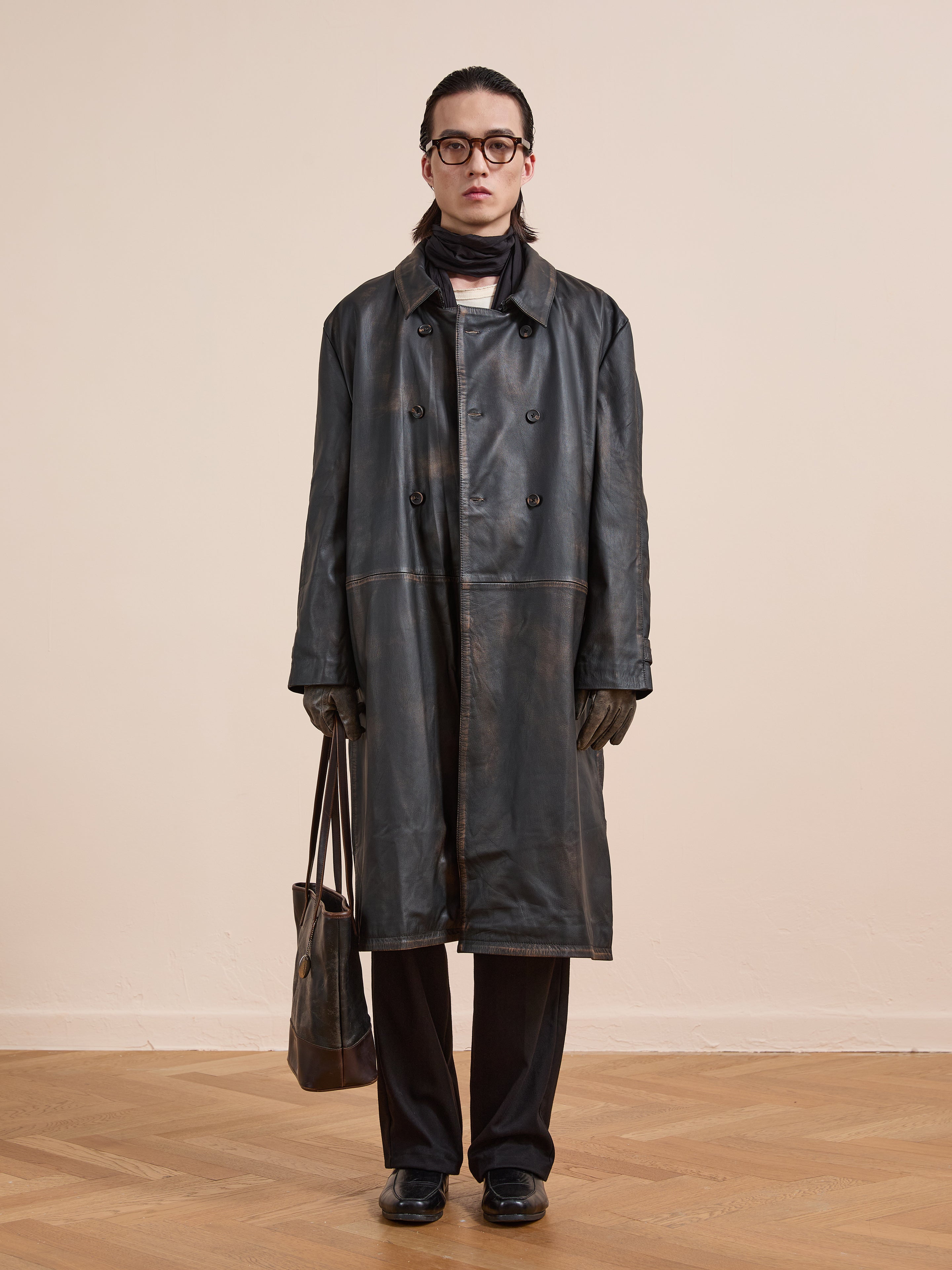 Individual wearing the FOUND Distressed Leather Trench Coat, black pants, gloves, glasses, and a scarf stands indoors on a wooden floor holding a leather bag.