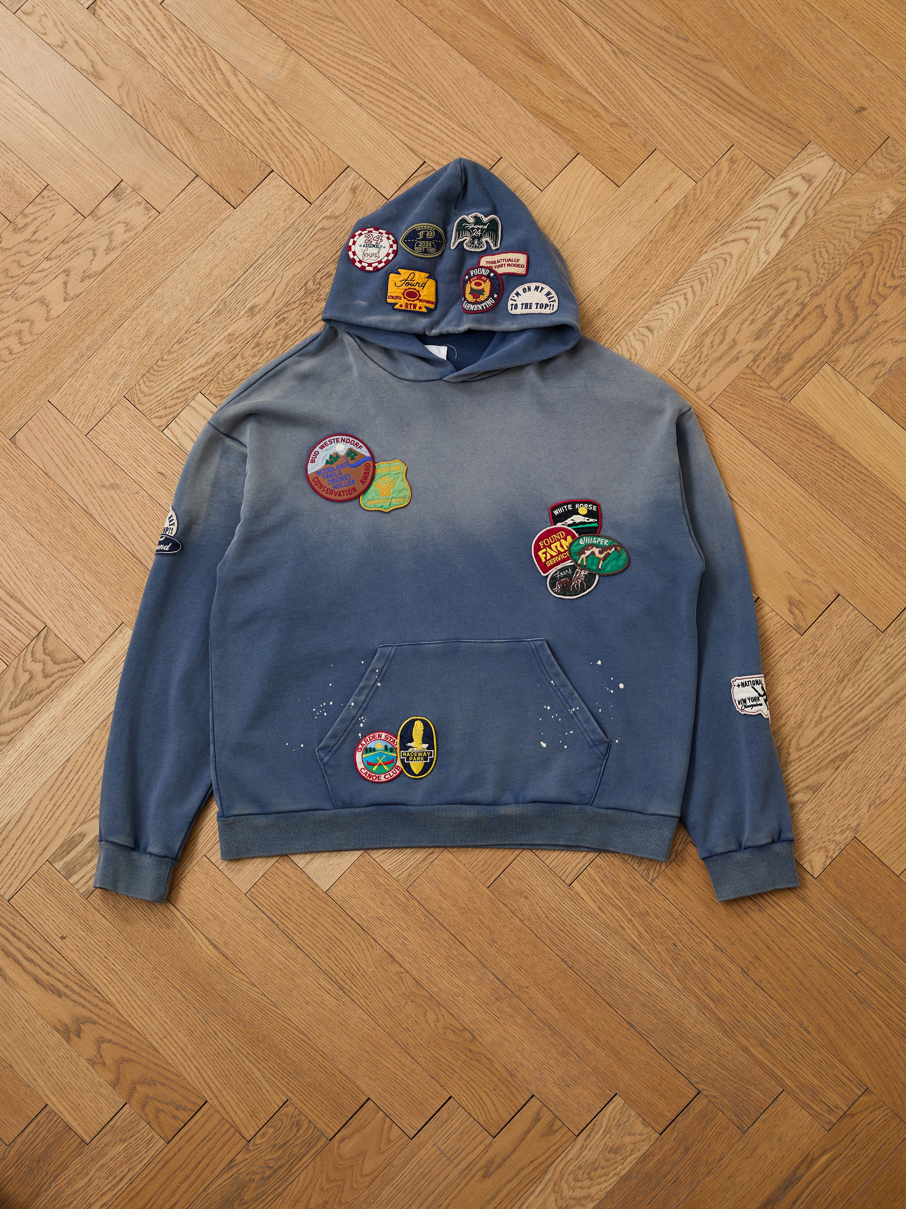 The FOUND Distressed Patch Hoodie in blue, made from soft French Terry cotton, showcases vibrant embroidered patches on the hood, chest, pocket, and sleeves—displayed flat on a herringbone wood floor.