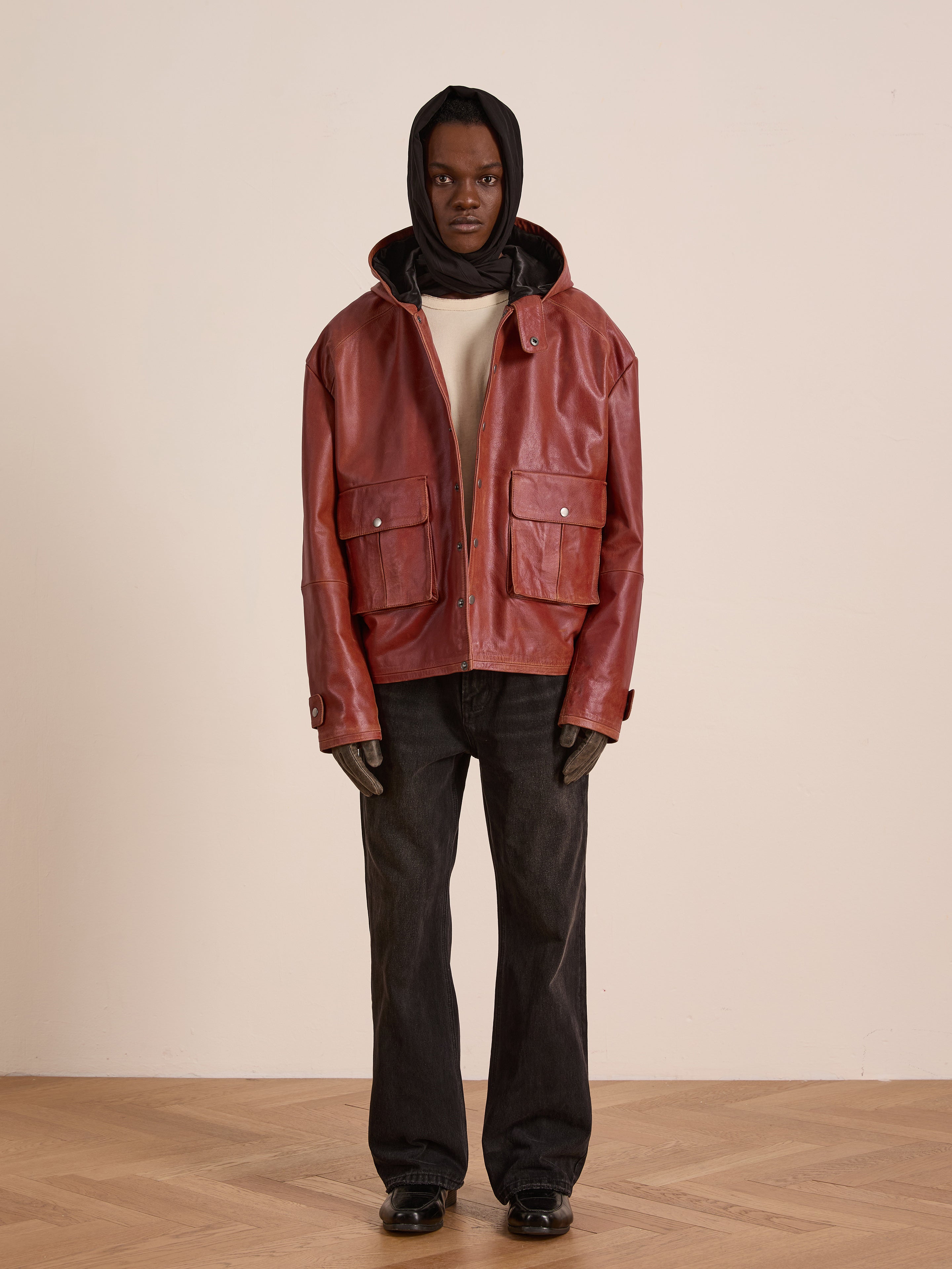 Indoors, a person wears the FOUND Distressed Leather Pocket Hoodie with dark jeans and black shoes against a plain light wall.