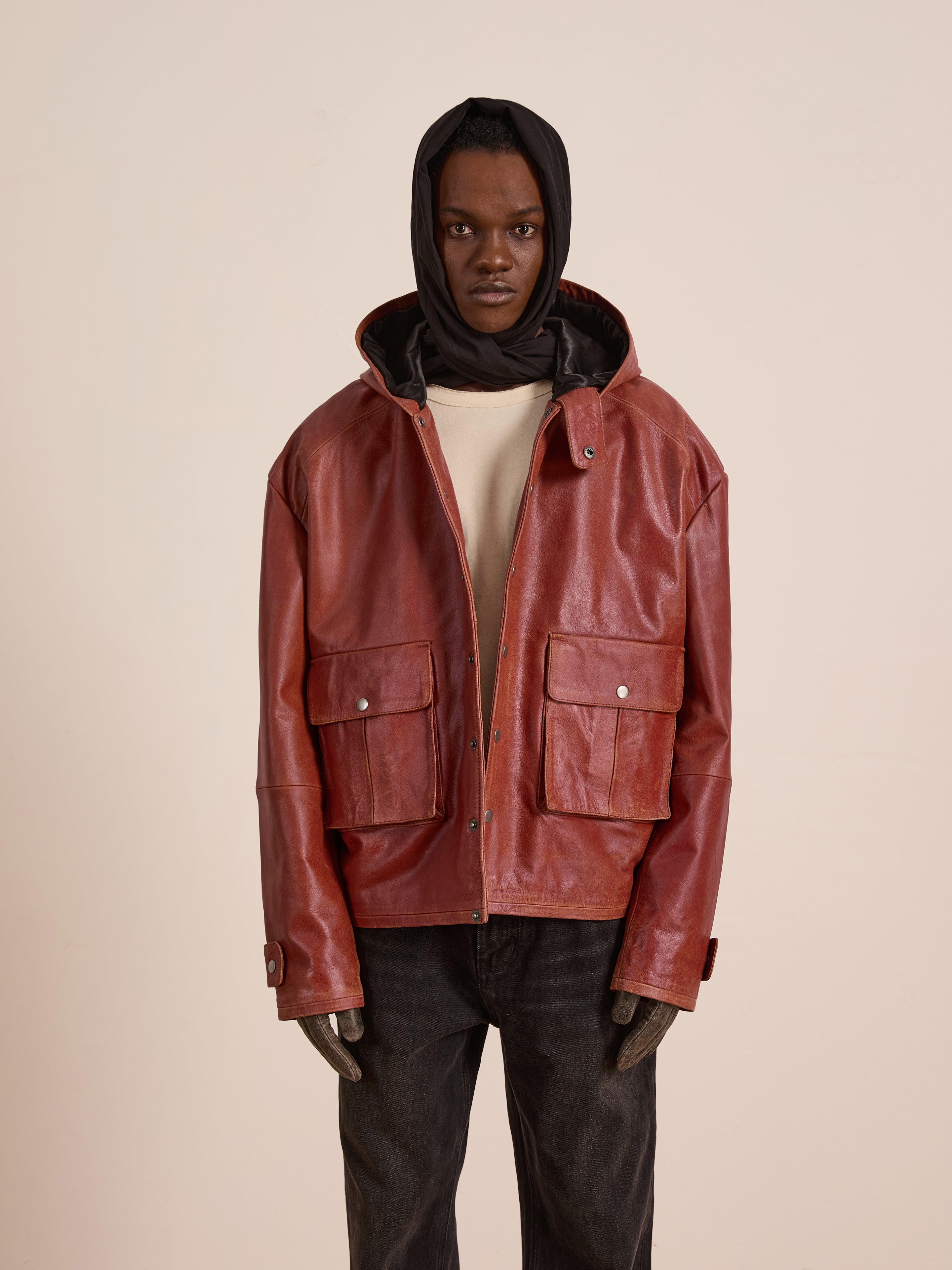A person wears the FOUND Distressed Leather Pocket Hoodie—a vintage-style red cowhide leather jacket with large front pockets—paired with a beige shirt, dark pants, and a black hooded head covering against a plain light background.