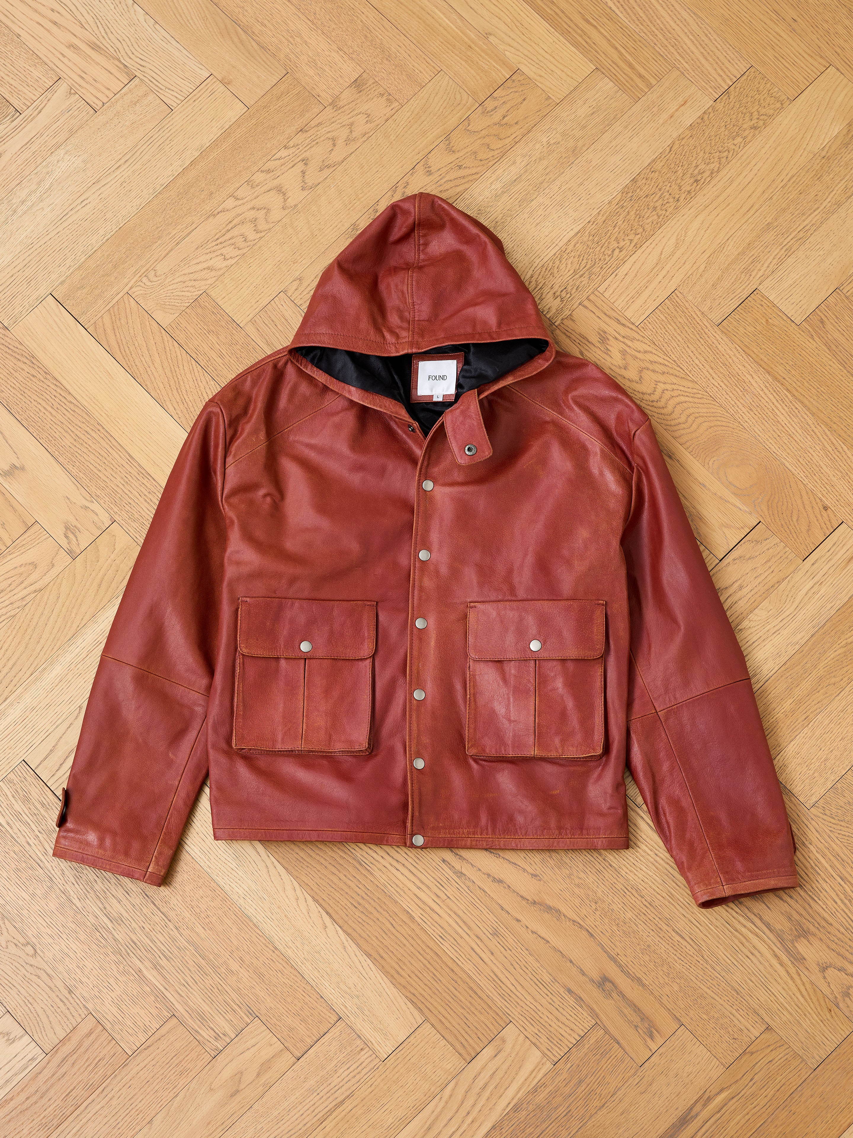 The FOUND Distressed Leather Pocket Hoodie, crafted from rich brown genuine cowhide, features snap buttons, two large front pockets, and a stylish hooded design—all laid flat on a light wooden parquet floor.