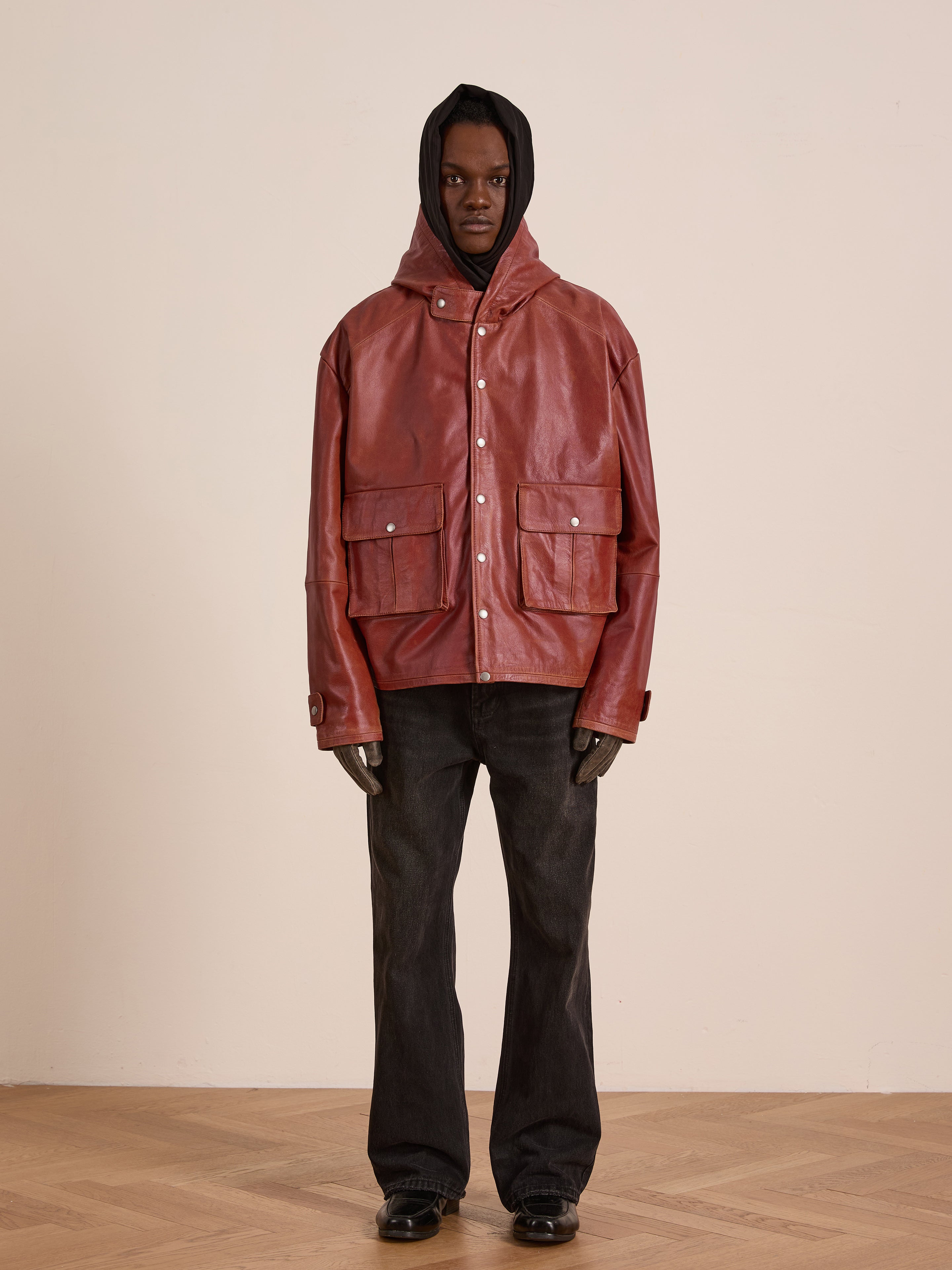 Indoors, a person wears the FOUND Distressed Leather Pocket Hoodie—vintage red cowhide with large front pockets—paired with black pants and shoes, standing against a plain light-colored wall.