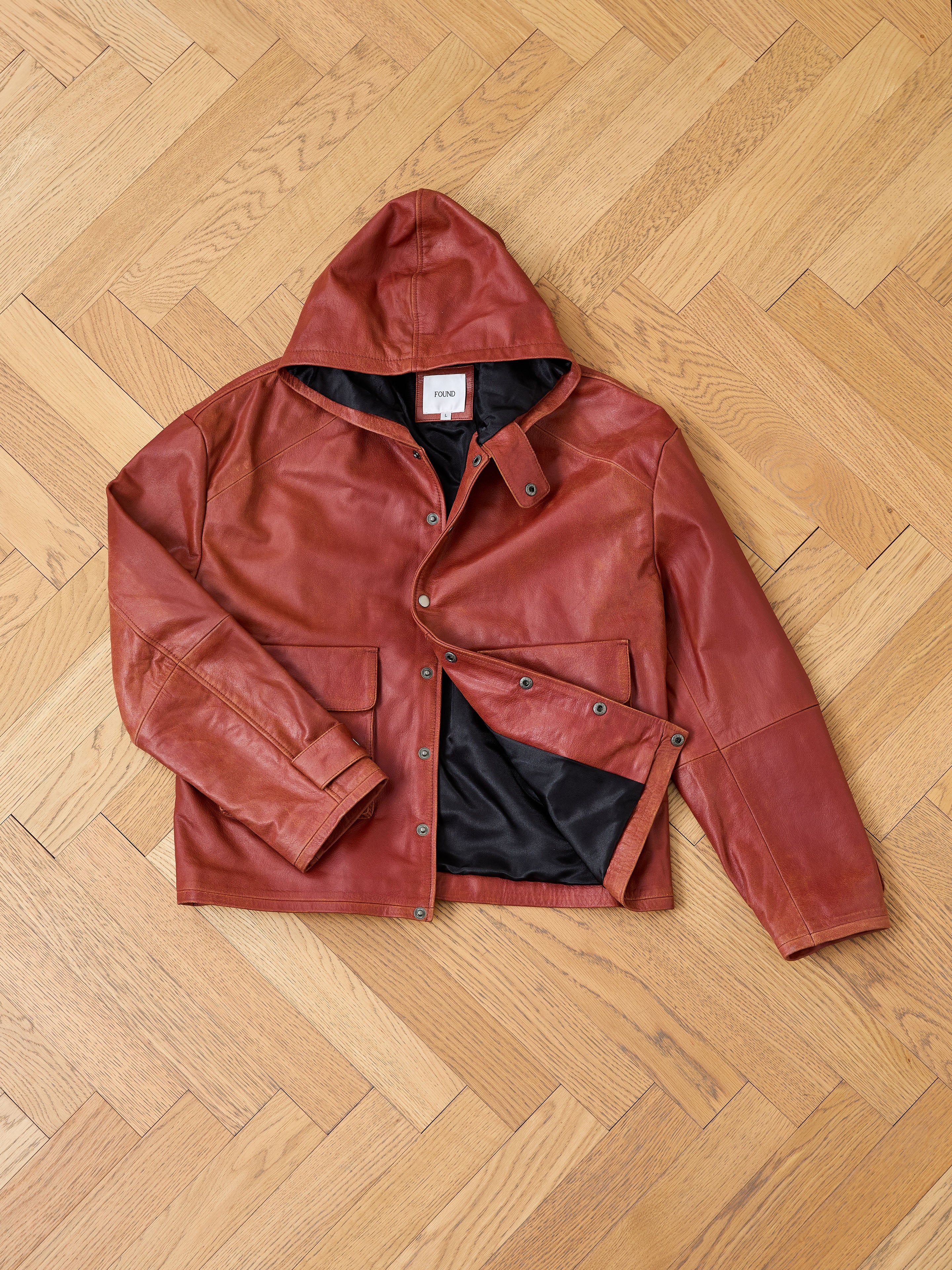 The FOUND Distressed Leather Pocket Hoodie, crafted from rich brown genuine cowhide leather with a black inner lining, is laid flat on a light wooden parquet floor, highlighting its stylish hooded design.