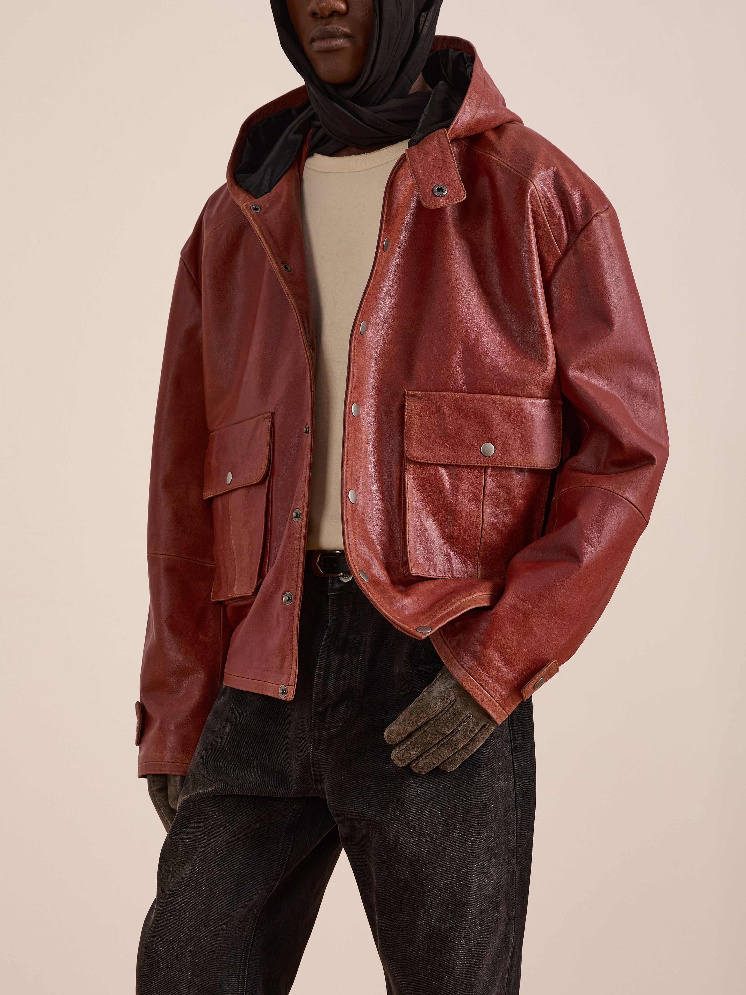 Model wears the FOUND Distressed Leather Pocket Hoodie in vintage red cowhide leather with large front pockets, paired with a beige shirt, dark jeans, gloves, and a black head covering against a plain background.