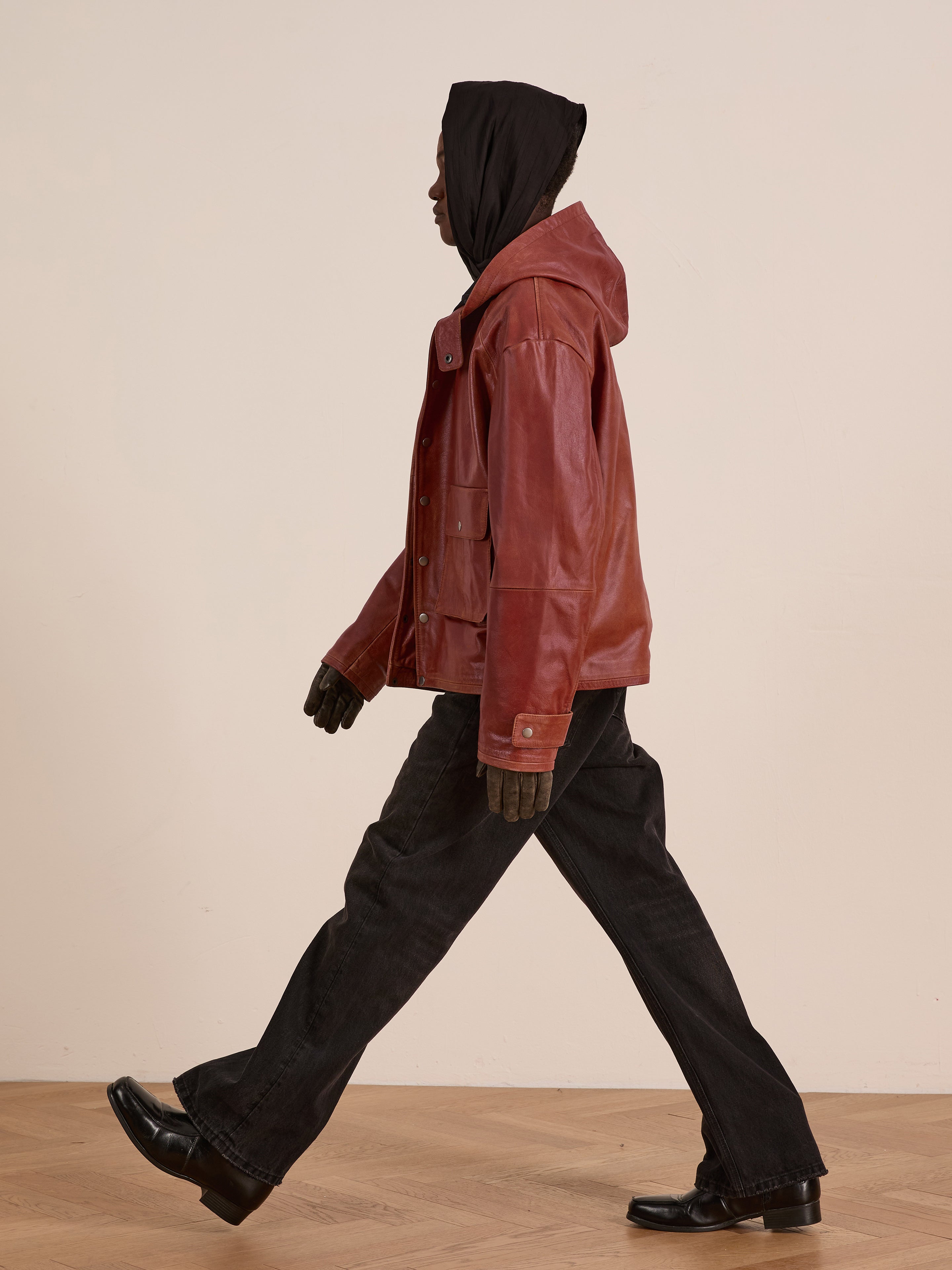 Wearing a FOUND Distressed Leather Pocket Hoodie, dark jeans, gloves, and black shoes, a person walks indoors against a plain background.