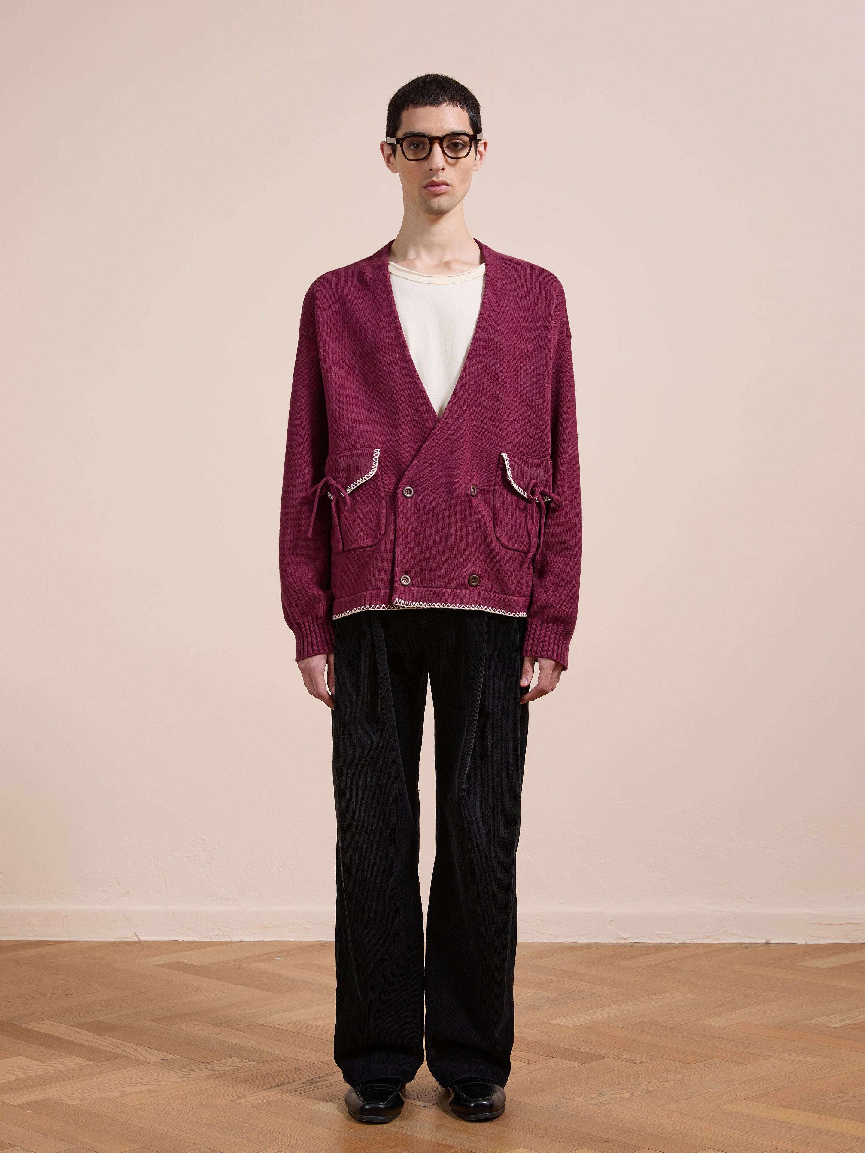 A person stands indoors against a plain beige background wearing the FOUND Double Breasted Bow Cardigan in burgundy with front pockets, layered over a white t-shirt, paired with black trousers, black shoes, and glasses.