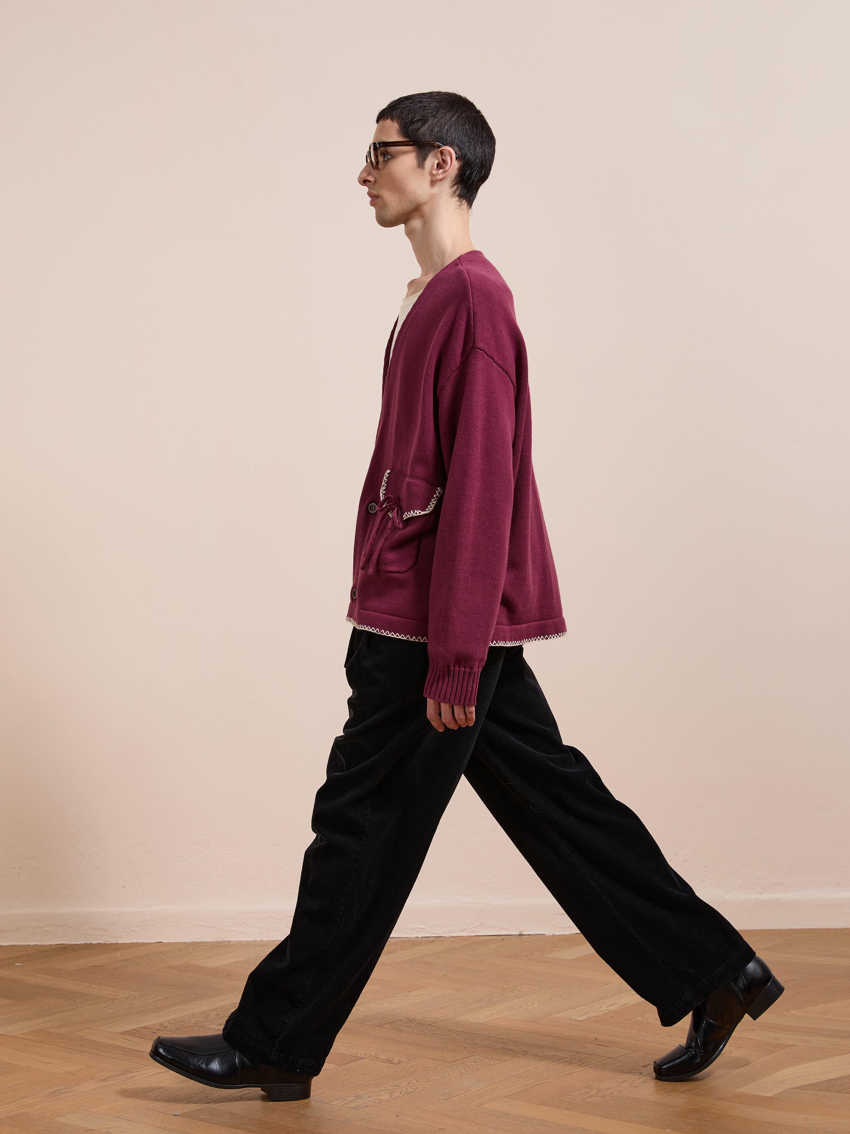 A person in the FOUND Double Breasted Bow Cardigan, black pants, and black shoes walks across a wooden floor against a plain beige wall.