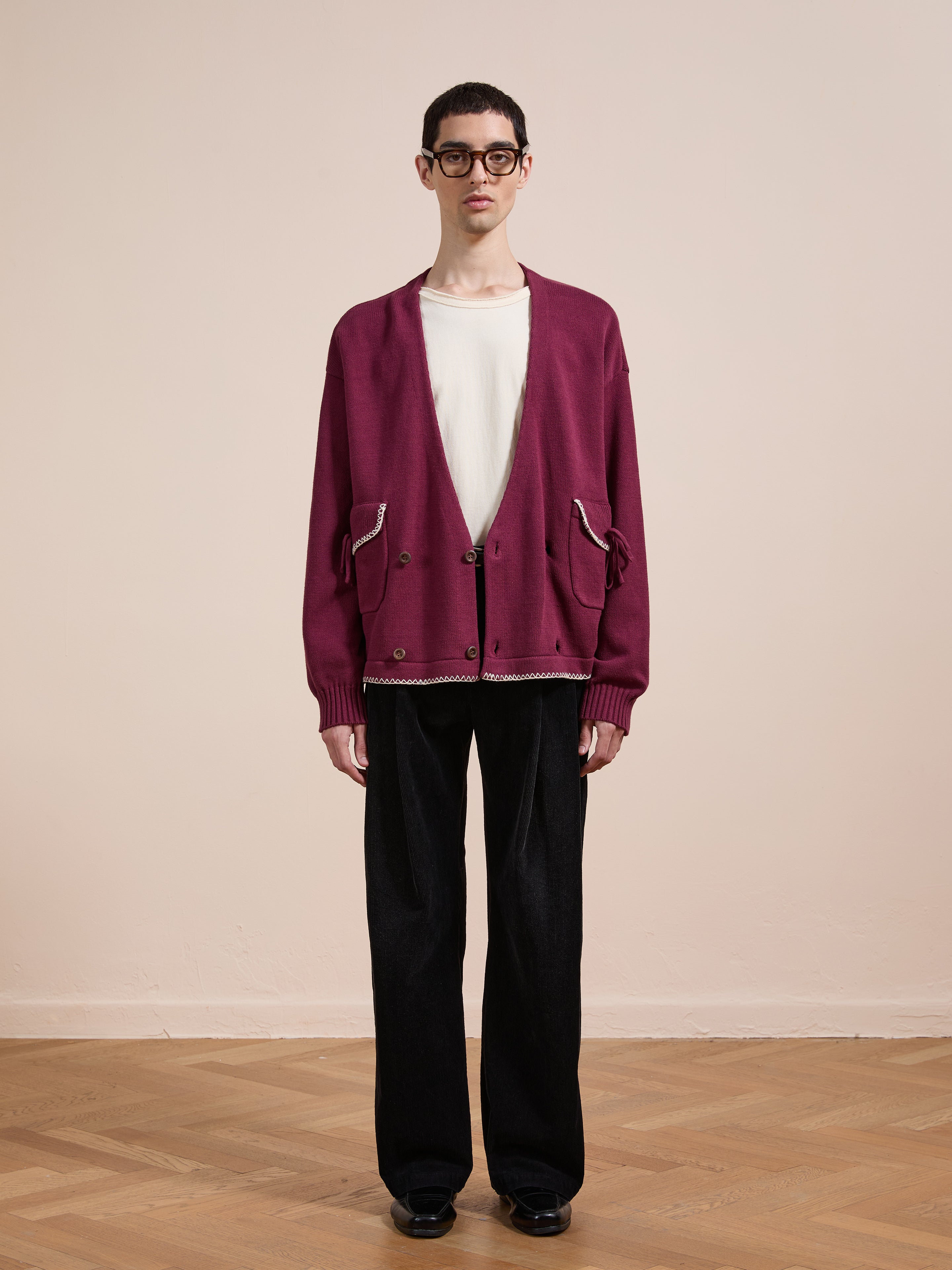 A person stands indoors on a wooden floor, wearing black pants, a white t-shirt, the FOUND Double Breasted Bow Cardigan in maroon, black shoes, and glasses, facing the camera against a plain beige wall.