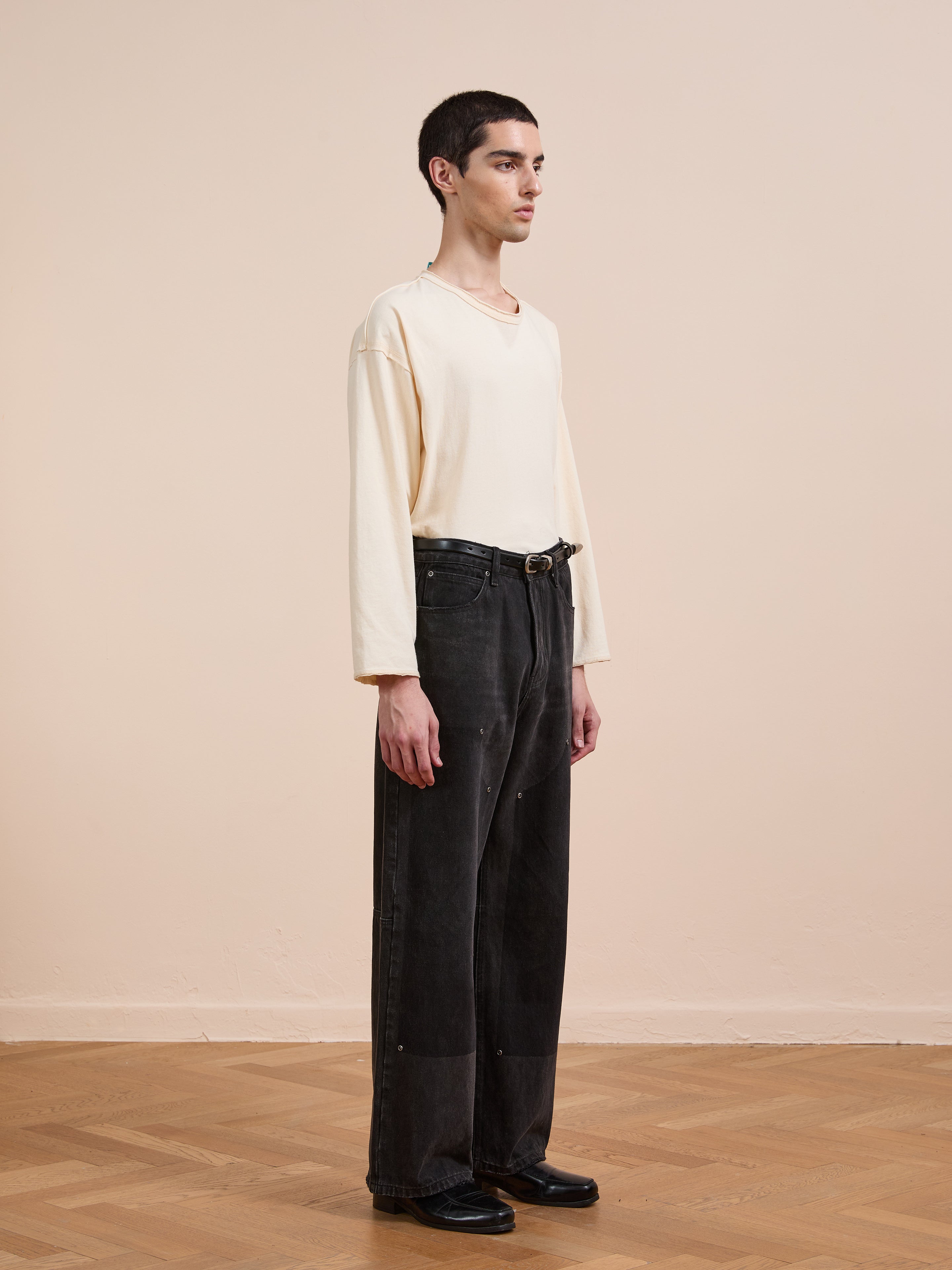 A person stands indoors on a wooden floor, wearing a loose cream long-sleeve shirt, black FOUND Double Knee Carpenter Jeans with a belt, and black shoes, facing left in a neutral pose.
