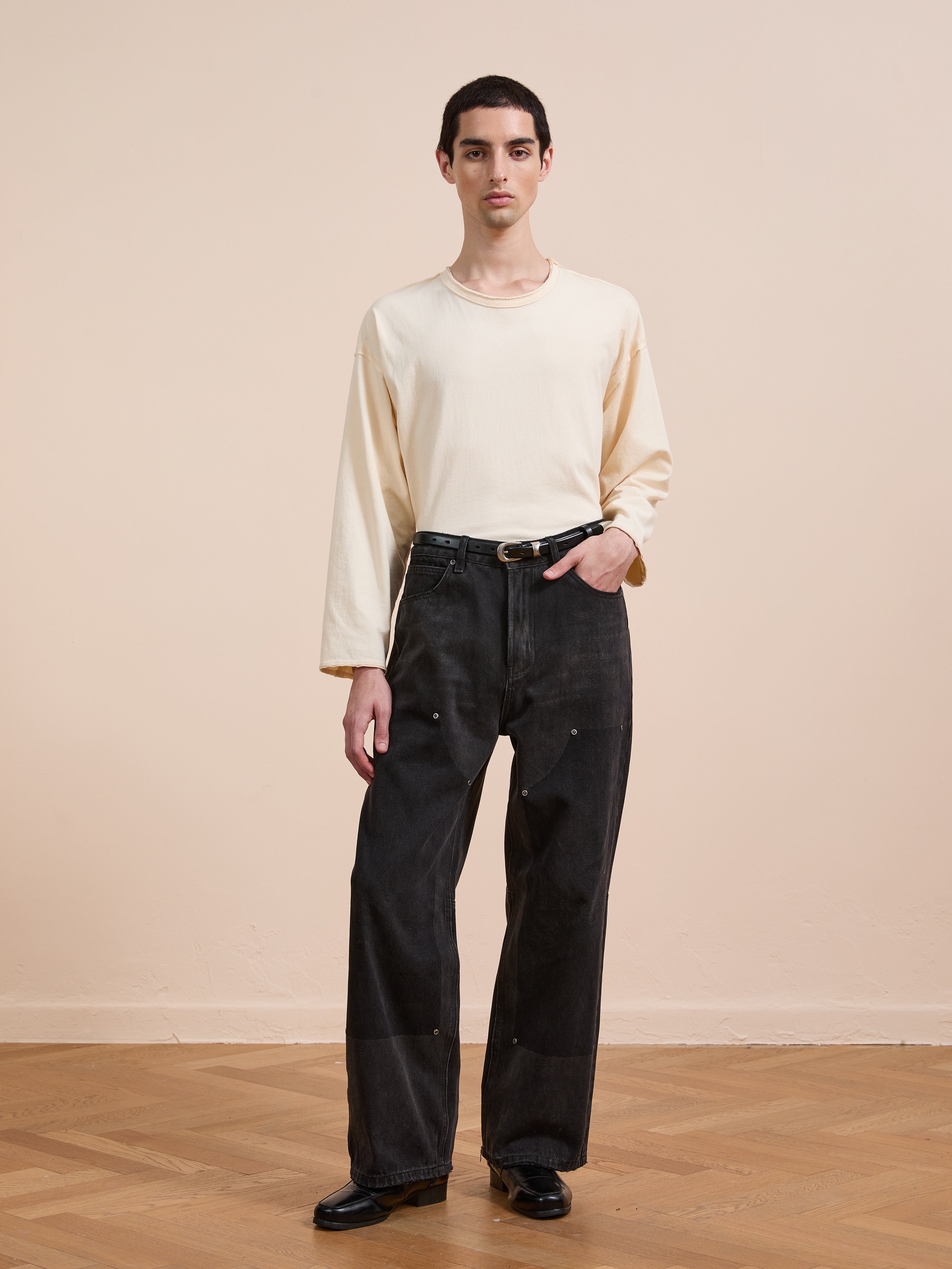 A person stands indoors on a wooden floor against a beige wall, wearing a long-sleeve cream shirt, black belt, black shoes, and FOUND Double Knee Carpenter Jeans with a relaxed wide-leg fit.