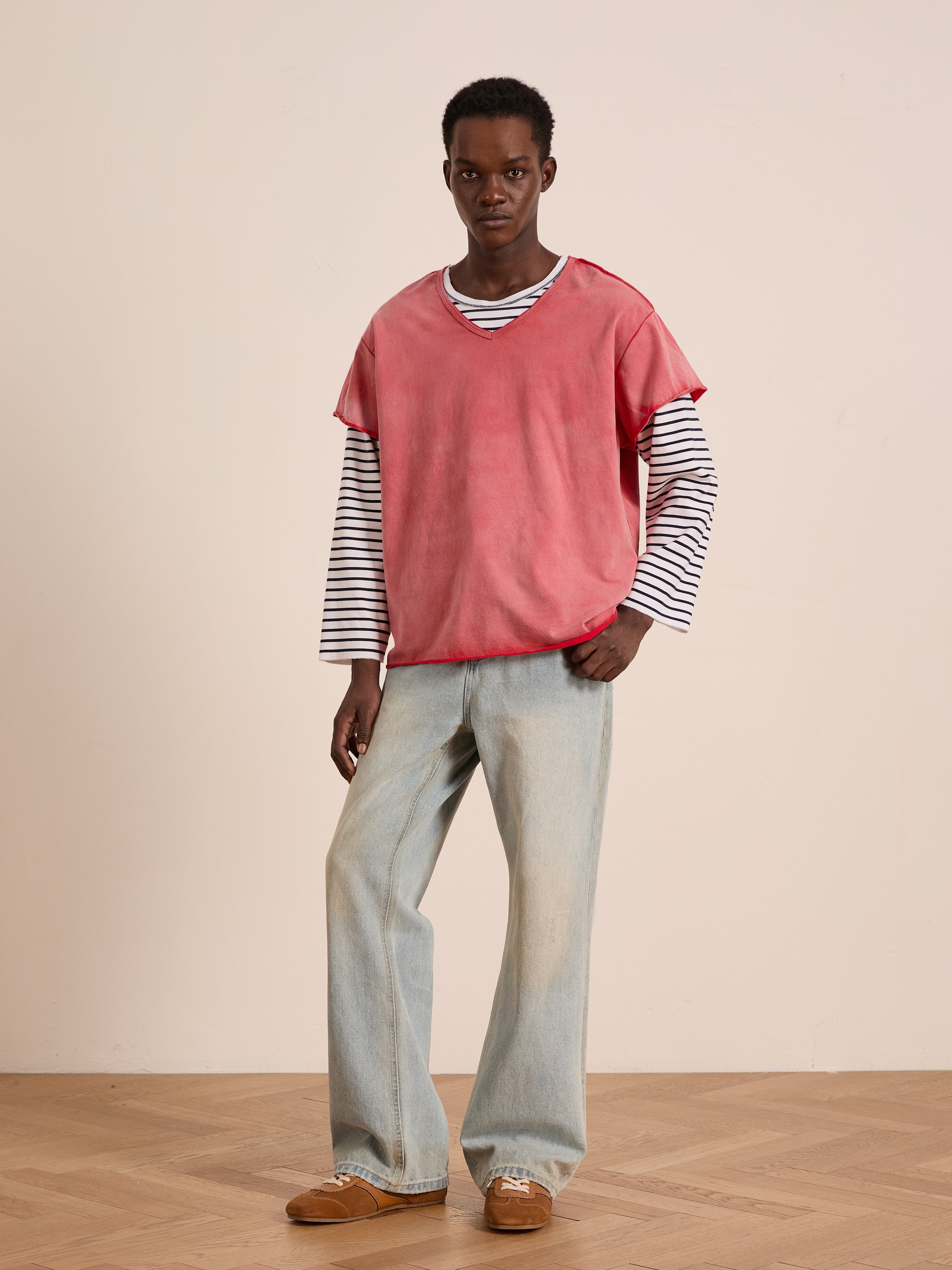 A person models FOUND’s Double Layer LS Tee—red short-sleeve over black and white stripes—in a relaxed unisex fit, paired with light blue jeans and brown shoes. The look stands out against a plain neutral background.