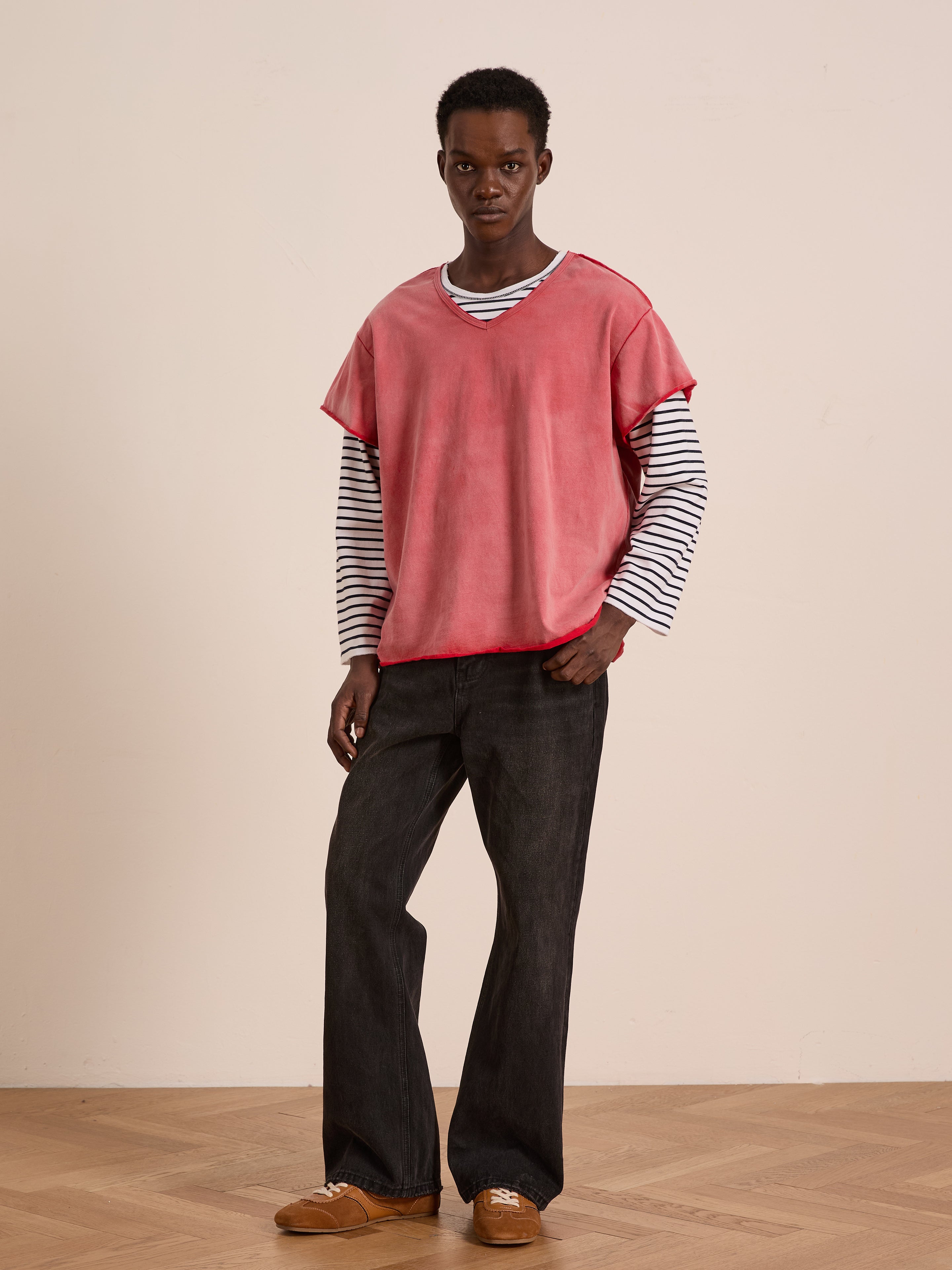 Indoors, a person wears FOUND’s Double Layer LS Tee in sunfade wash red over black and white stripes, styled with dark wide-leg pants and brown shoes on a wooden floor.