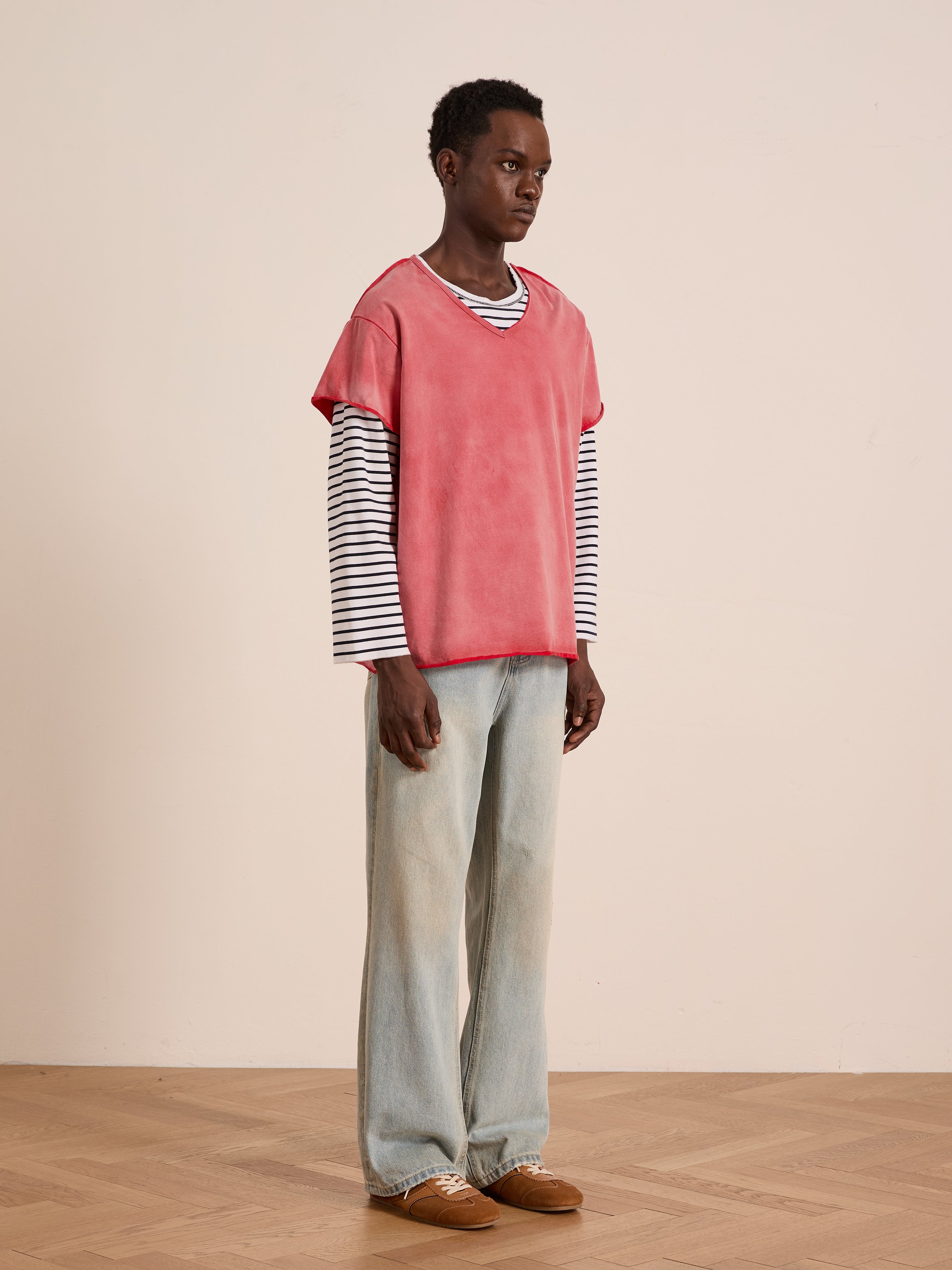 A person wears FOUND’s Double Layer LS Tee—red short sleeve over black and white stripes—with loose light blue jeans and brown shoes, showcasing a unisex relaxed fit in front of a plain beige background.