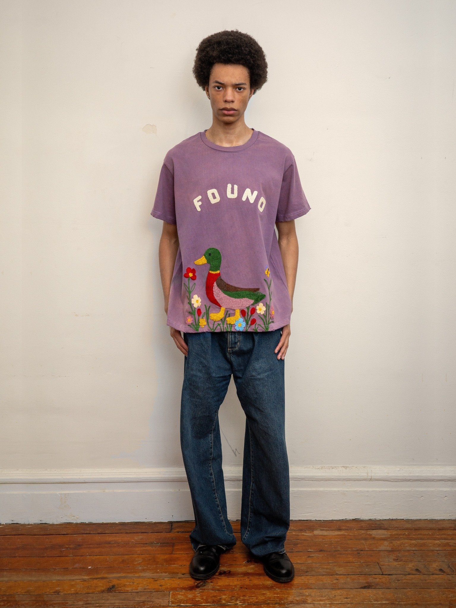 A person indoors wears the FOUND Embroidered Wildlife Tee featuring a purple color, chenille duck graphic, and "FOUND" text, paired with blue jeans and black shoes, standing against a plain wall and wooden floor.