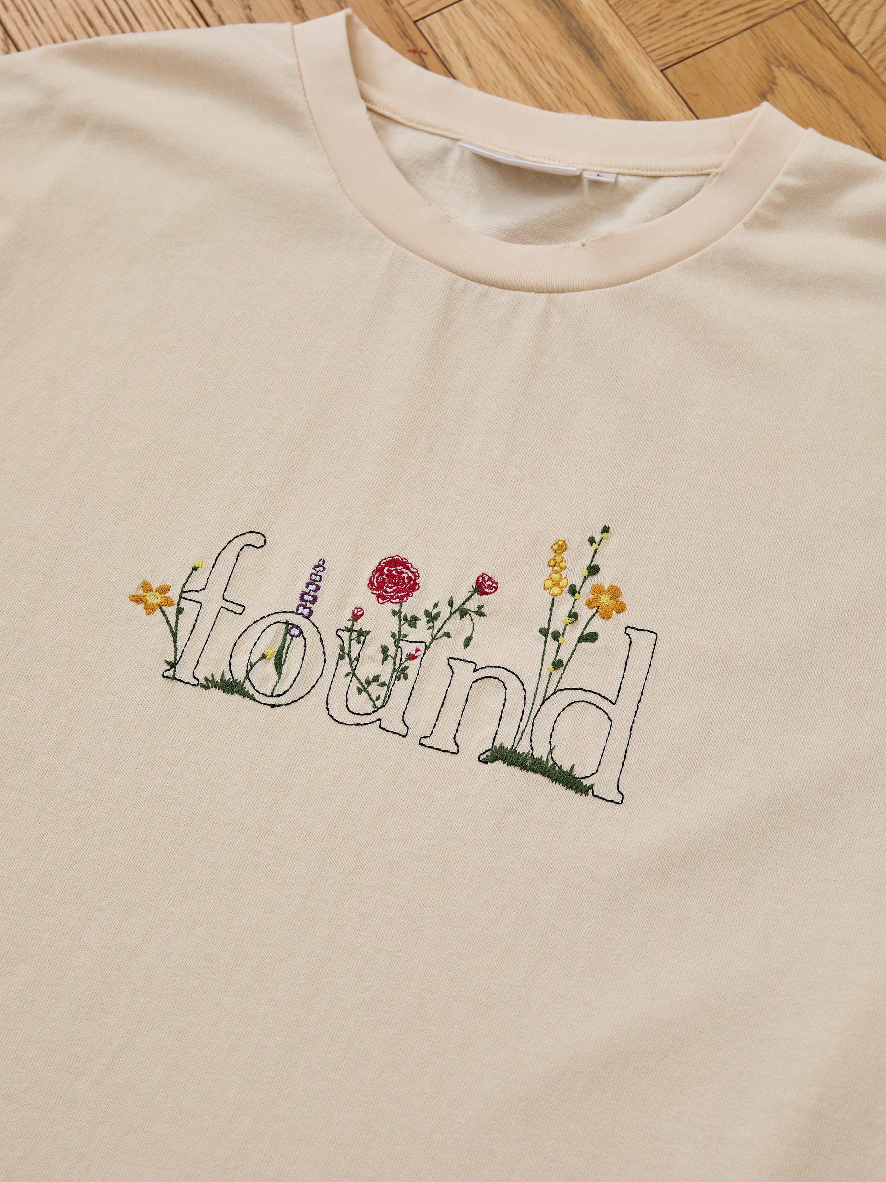 The FOUND Logo Flower Tee in beige features "found" in outlined letters and embroidered floral accents, adding a vintage-inspired touch.